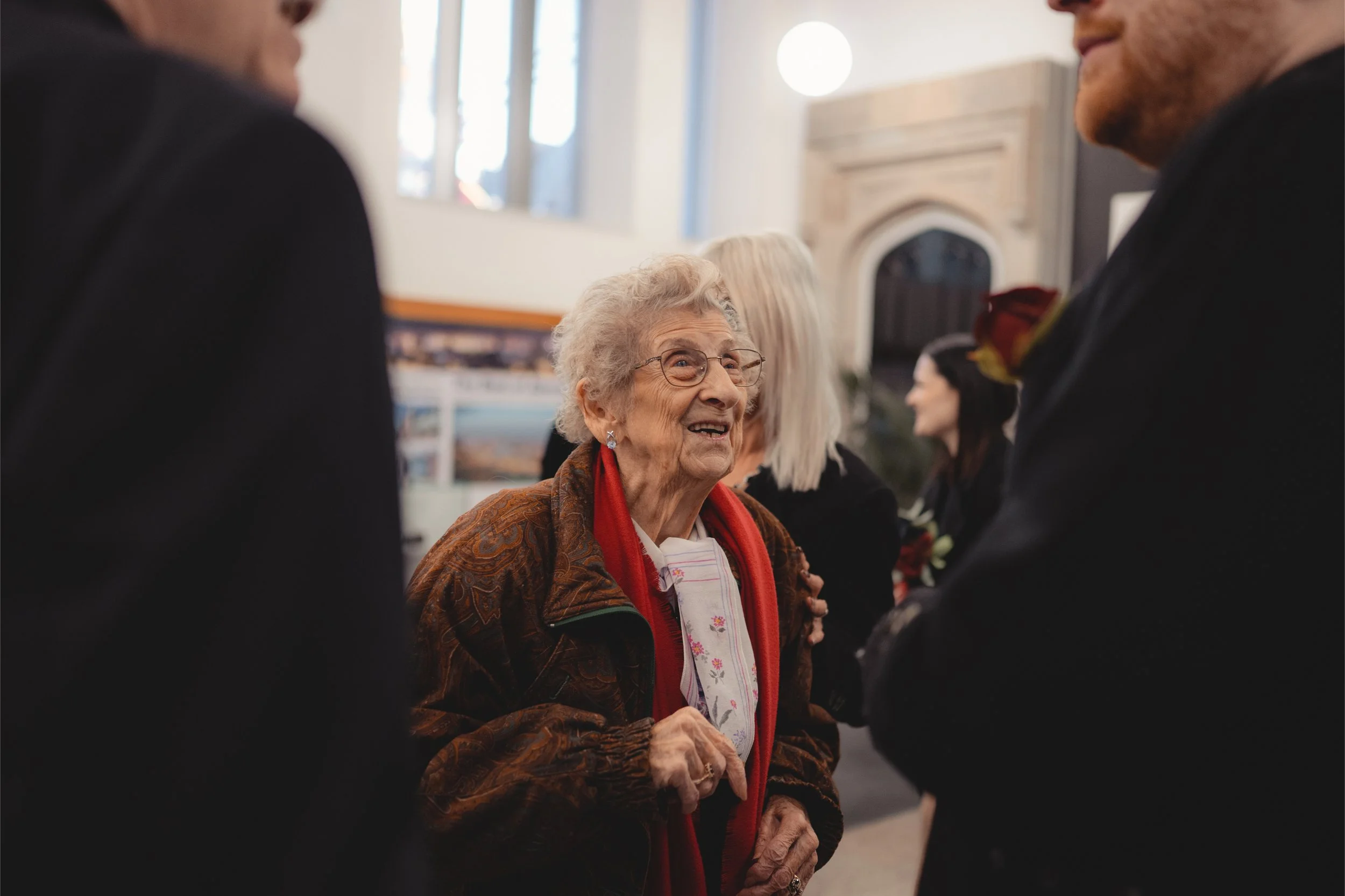 An elderly woman with gray hair, wearing glasses, a patterned brown jacket, and a red scarf, is smiling and engaging in conversation with two men dressed in formal attire during a social gathering indoors.