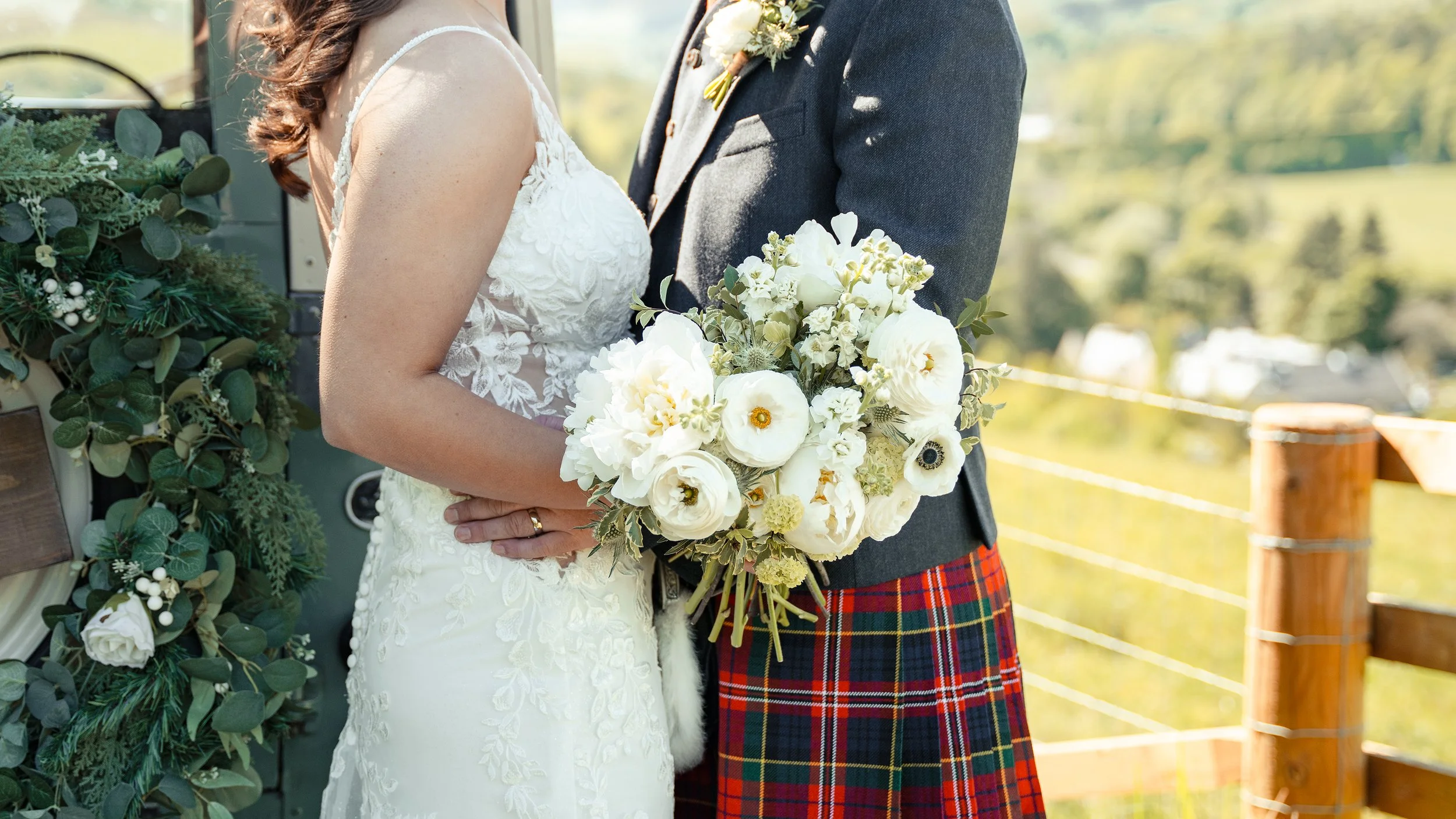Close-up of a bride and groom on their wedding day, holding a bouquet of white flowers, with the bride wearing a lace wedding dress and the groom dressed in a gray suit with red tartan kilt, outdoors with greenery in the background.