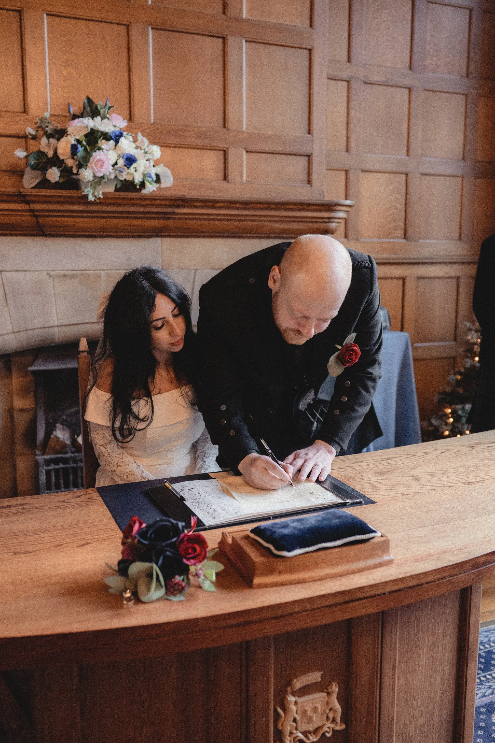 A woman in a white dress sitting at a wooden table while a man signs a document. The man has a bald head and is wearing a black jacket with a red rose pinned on it. There are flowers and a velvet pillow on the table, with a wooden crest carved into t