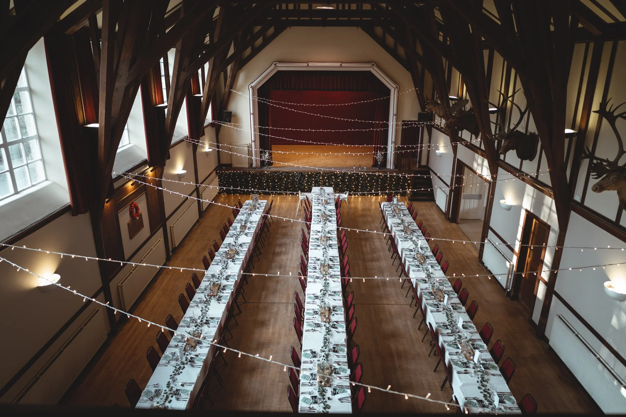 Long dining tables set with white tablecloths, glassware, and place settings in a large decorated hall with high wooden beams and string lights.