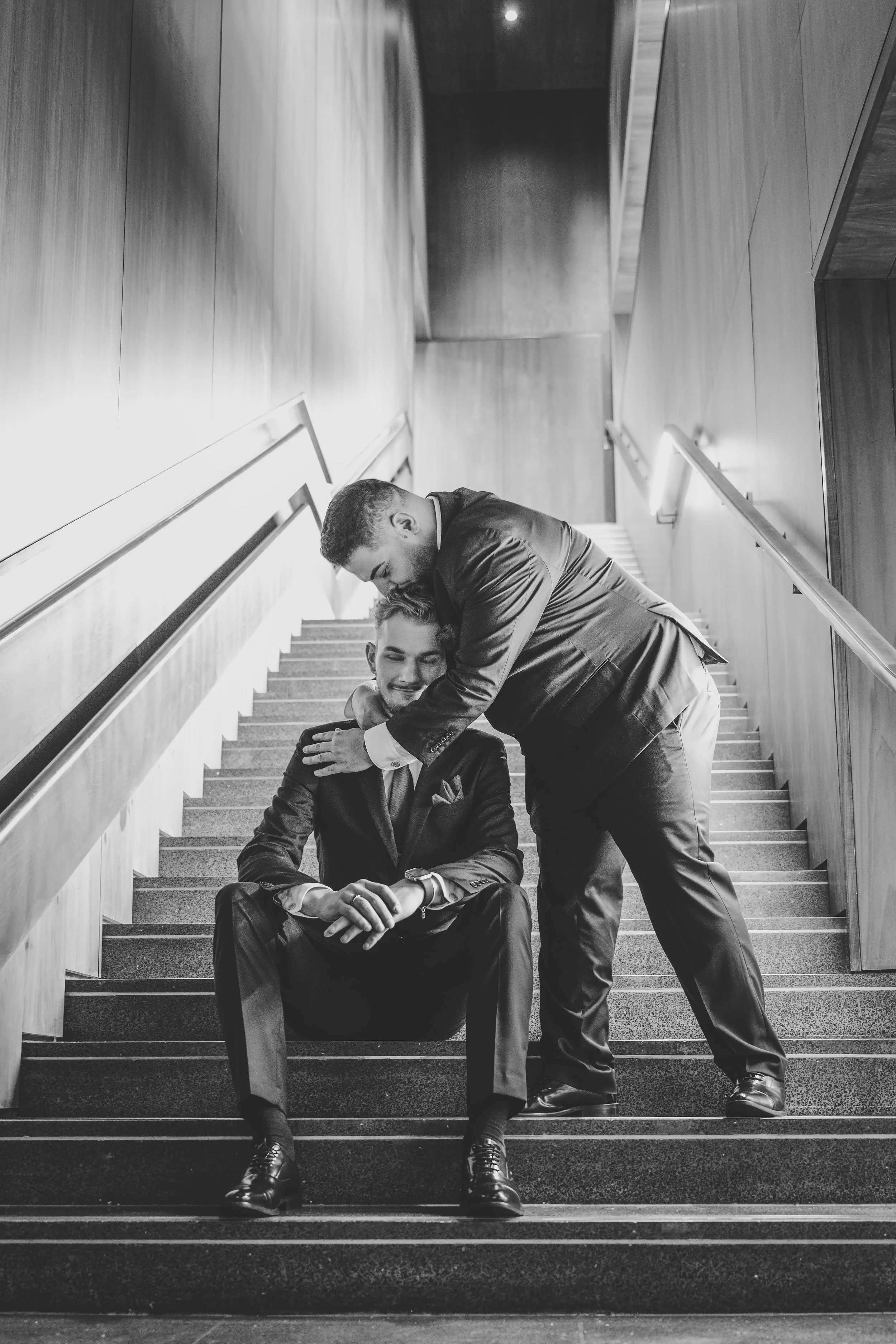 Two men in suits embracing on a staircase, one sitting and the other leaning down, in a black and white photo.