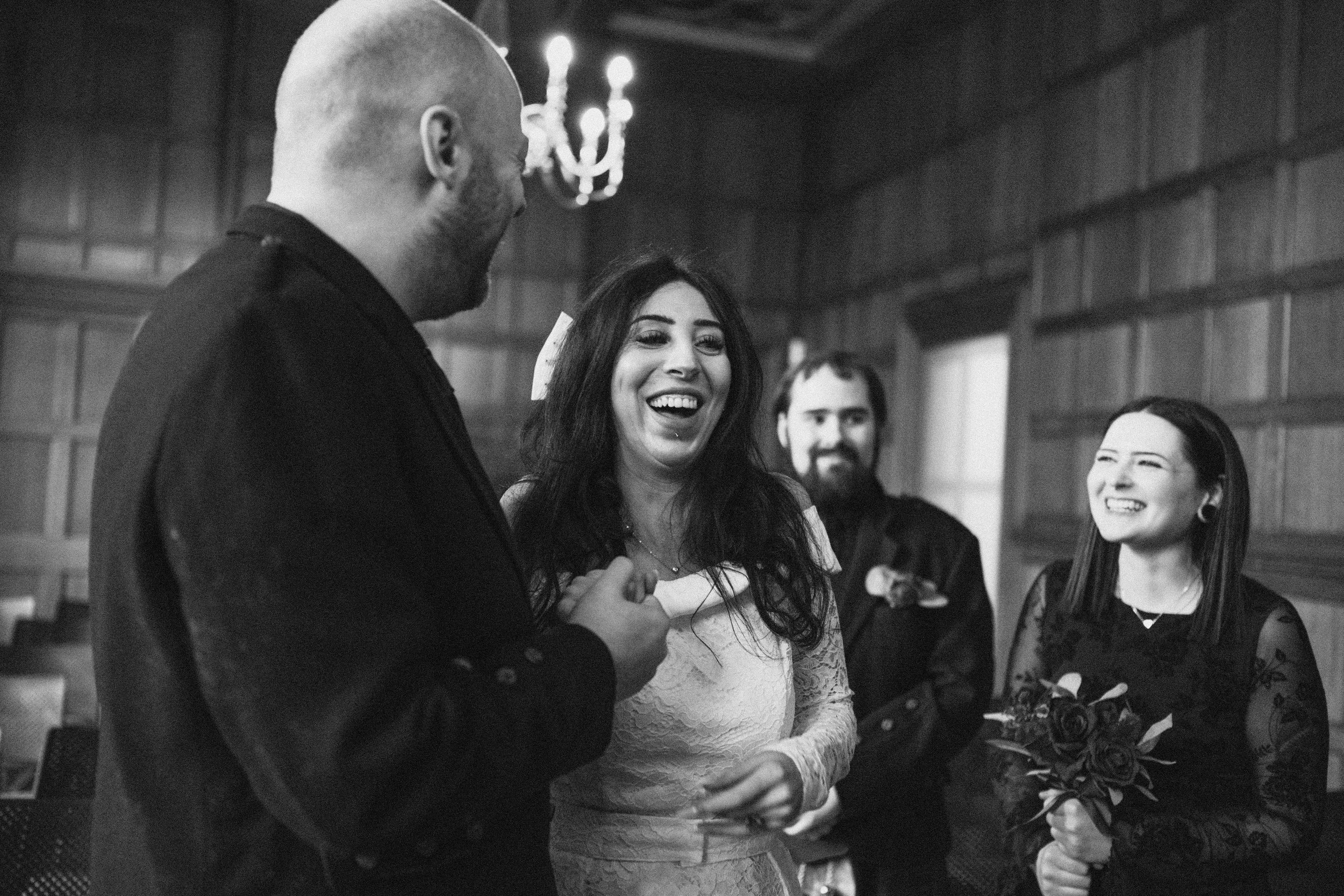 A group of four people smiling and talking in an indoor setting, with dark wood paneled walls and a chandelier visible in the background.