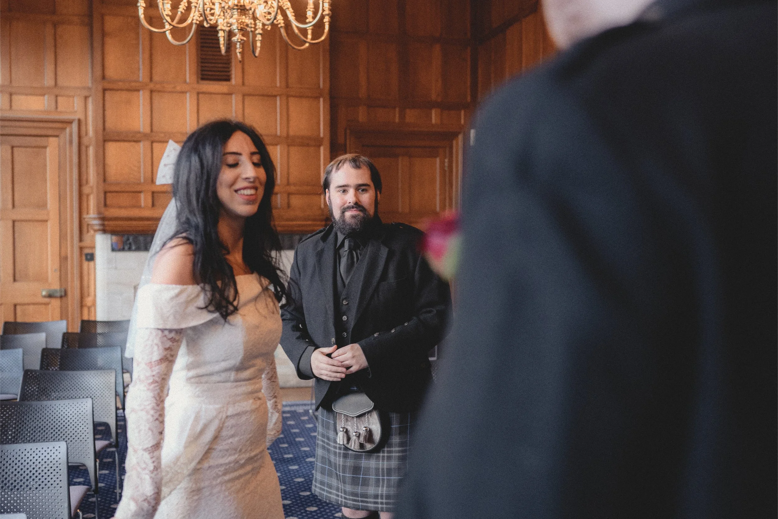 A bride in a white off-shoulder wedding dress smiling, with a man in a black jacket and tartan kilt beside her, inside a wood-paneled room with a chandelier.