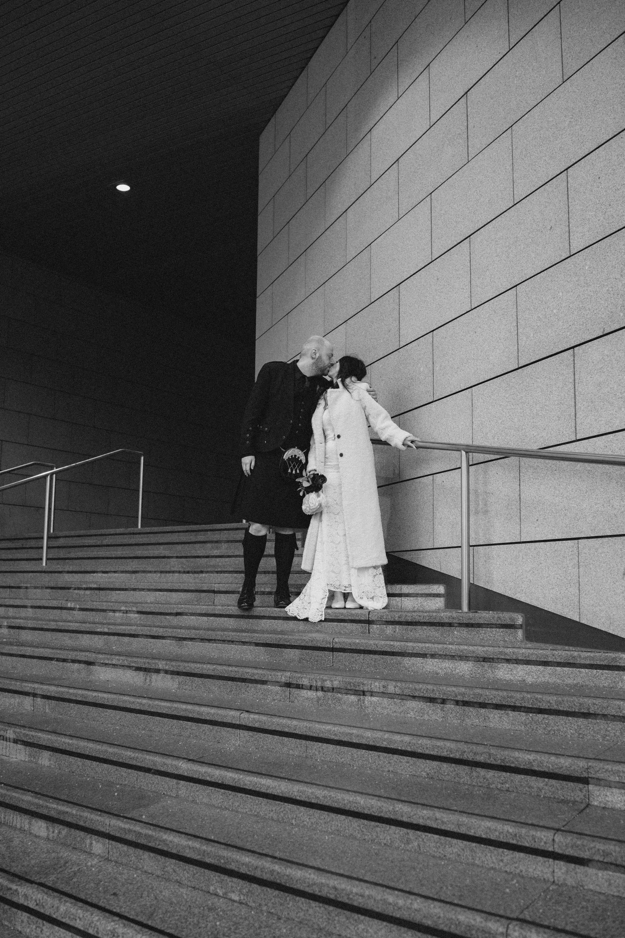 A black and white photo of a man and a woman standing on a staircase with a modern architectural background. The woman is wearing a long lace dress and coat, holding a bouquet. The man is dressed in traditional Scottish attire with a kilt and a jacke