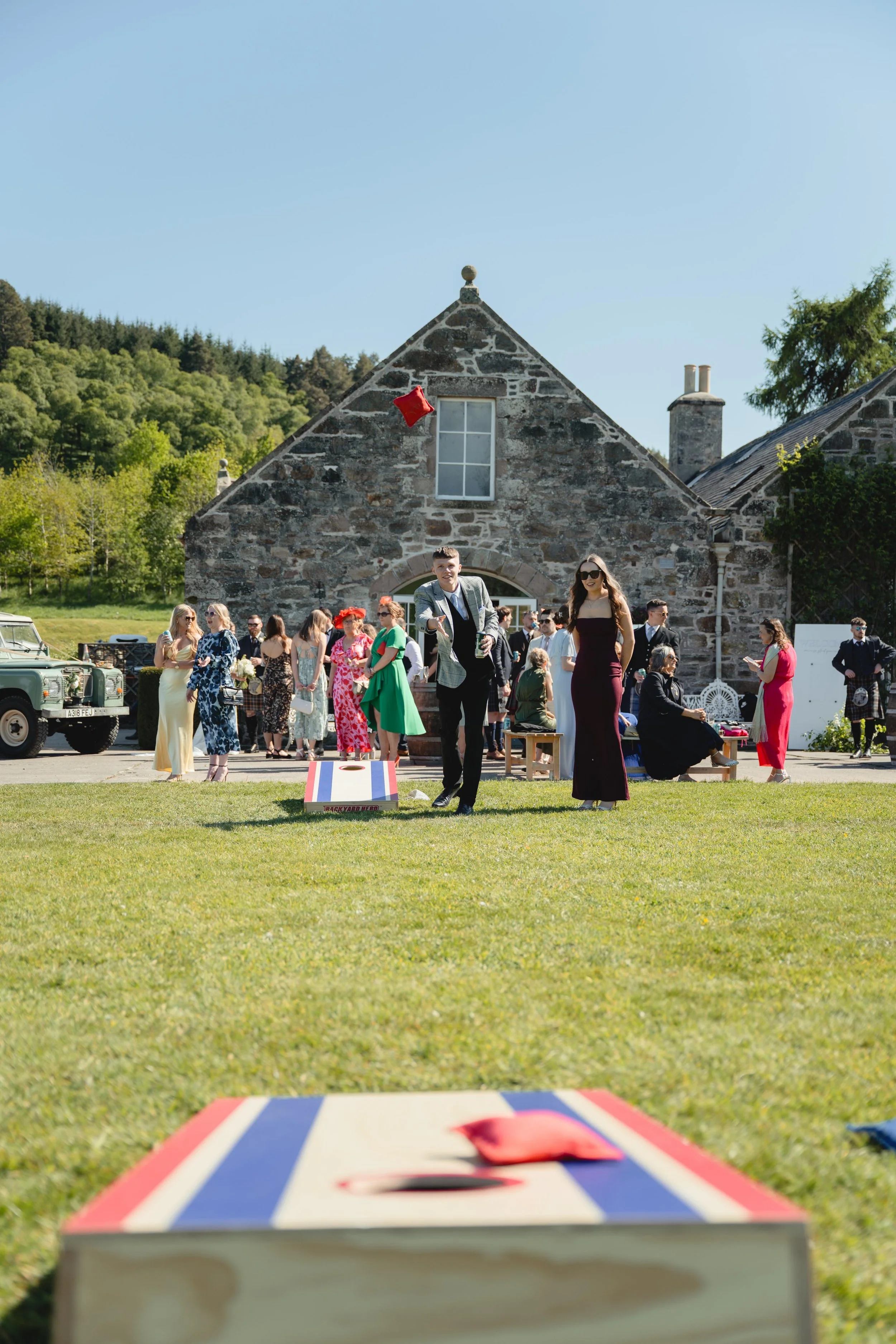 People playing cornhole at an outdoor wedding reception or celebration on a sunny day, with a stone building and green trees in the background.
