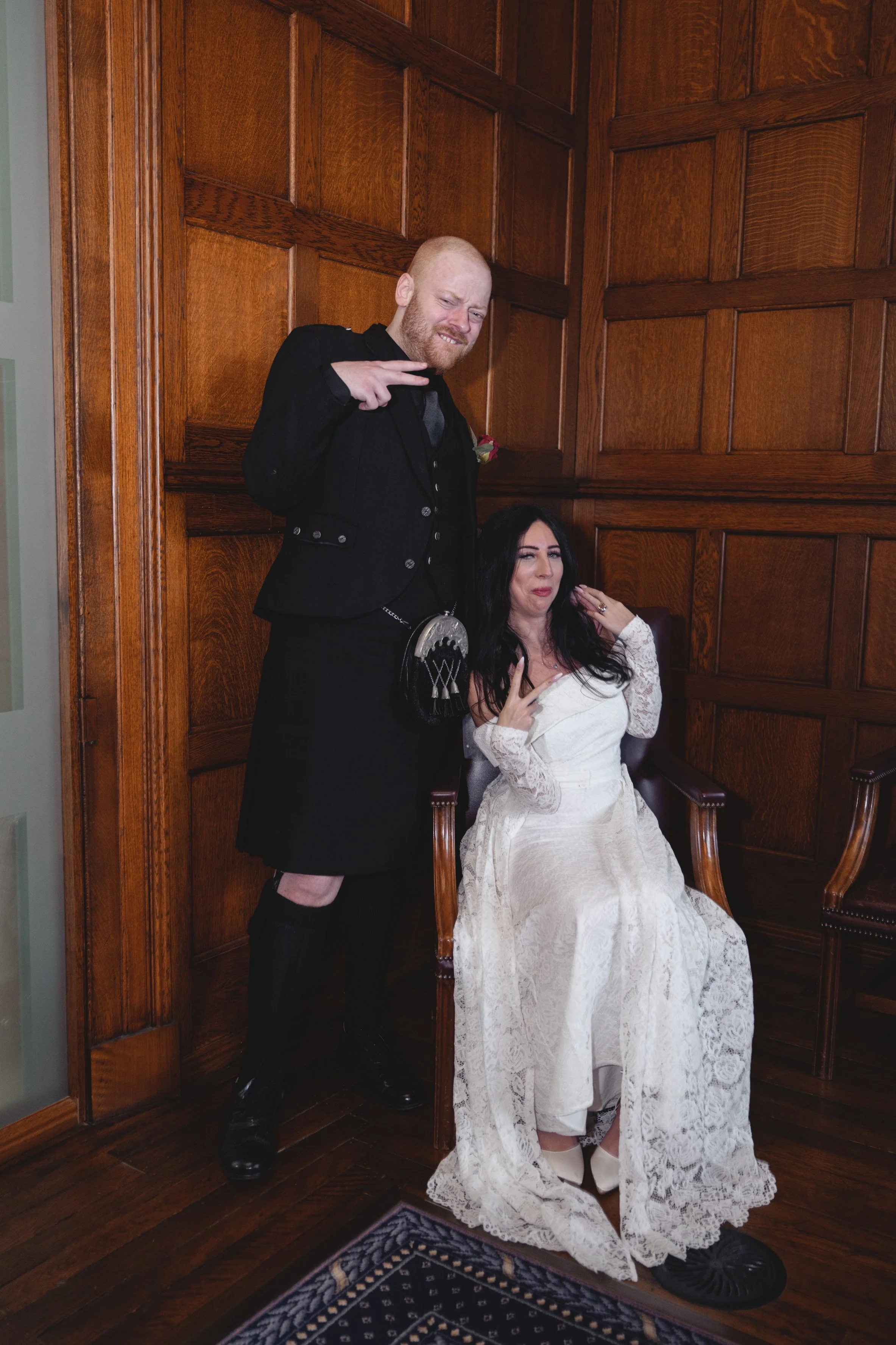 A man in a black kilt and jacket making a peace sign and a woman in a wedding dress sitting on a chair making a funny face.