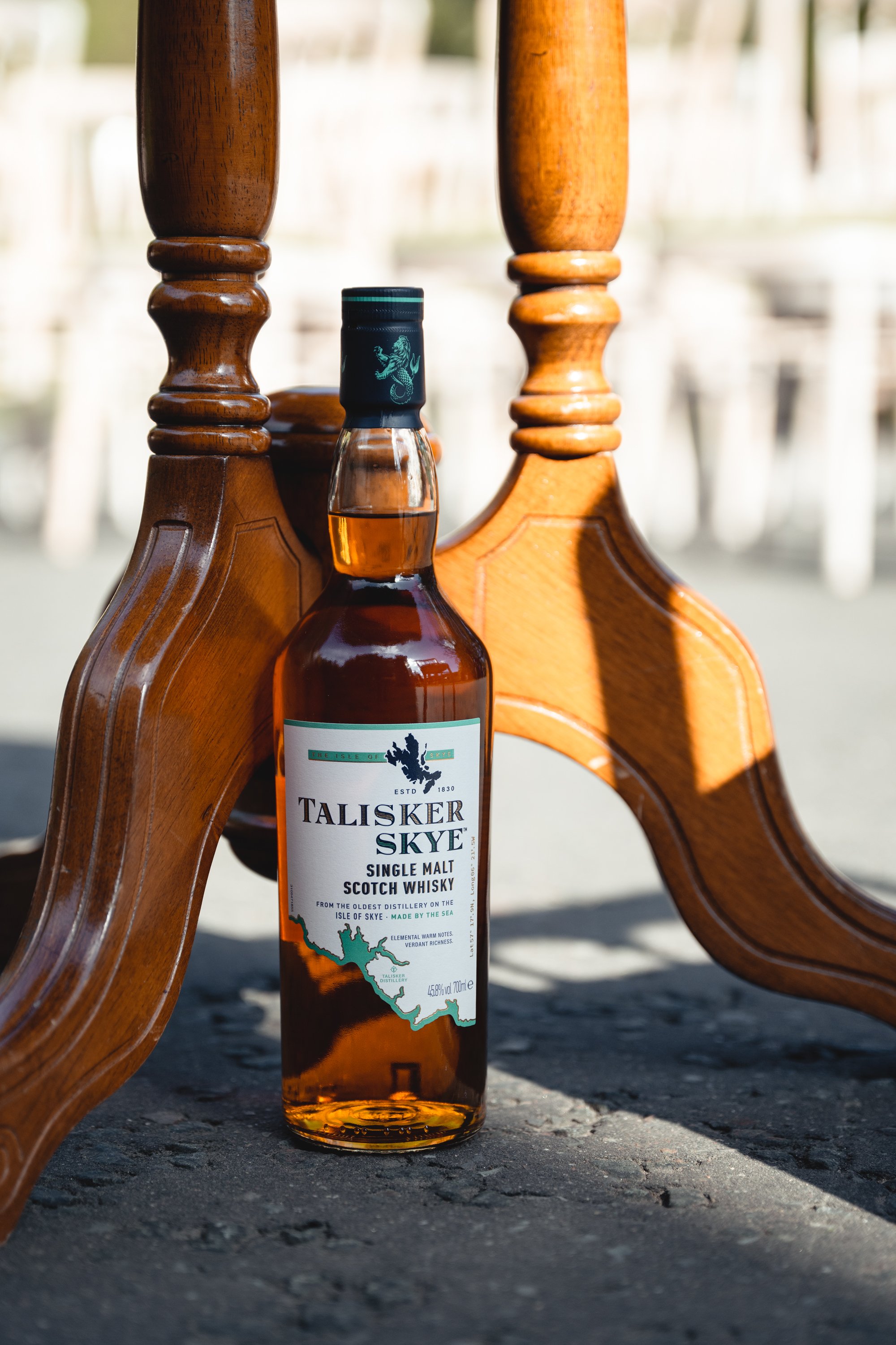 A bottle of Talisker Skye single malt Scotch whisky placed beneath a wooden chair or bench, with the focus on the bottle's label and the chair's wooden legs. The scene appears outdoors with natural lighting.