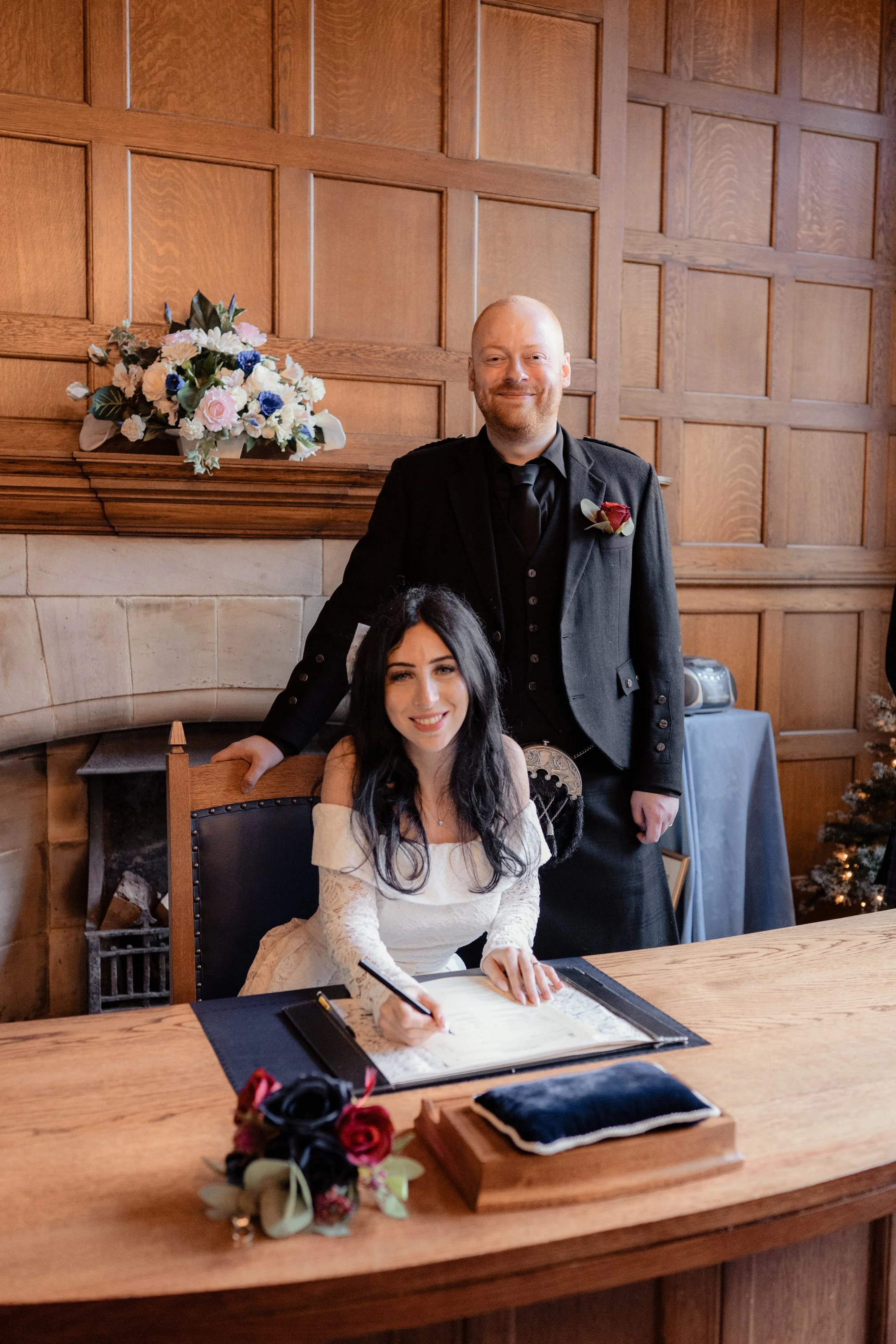 A woman wearing a white wedding dress signing a document at a table, with a man in a black suit standing behind her, both smiling, in a wood-paneled room decorated for Christmas with a small Christmas tree and flowers.