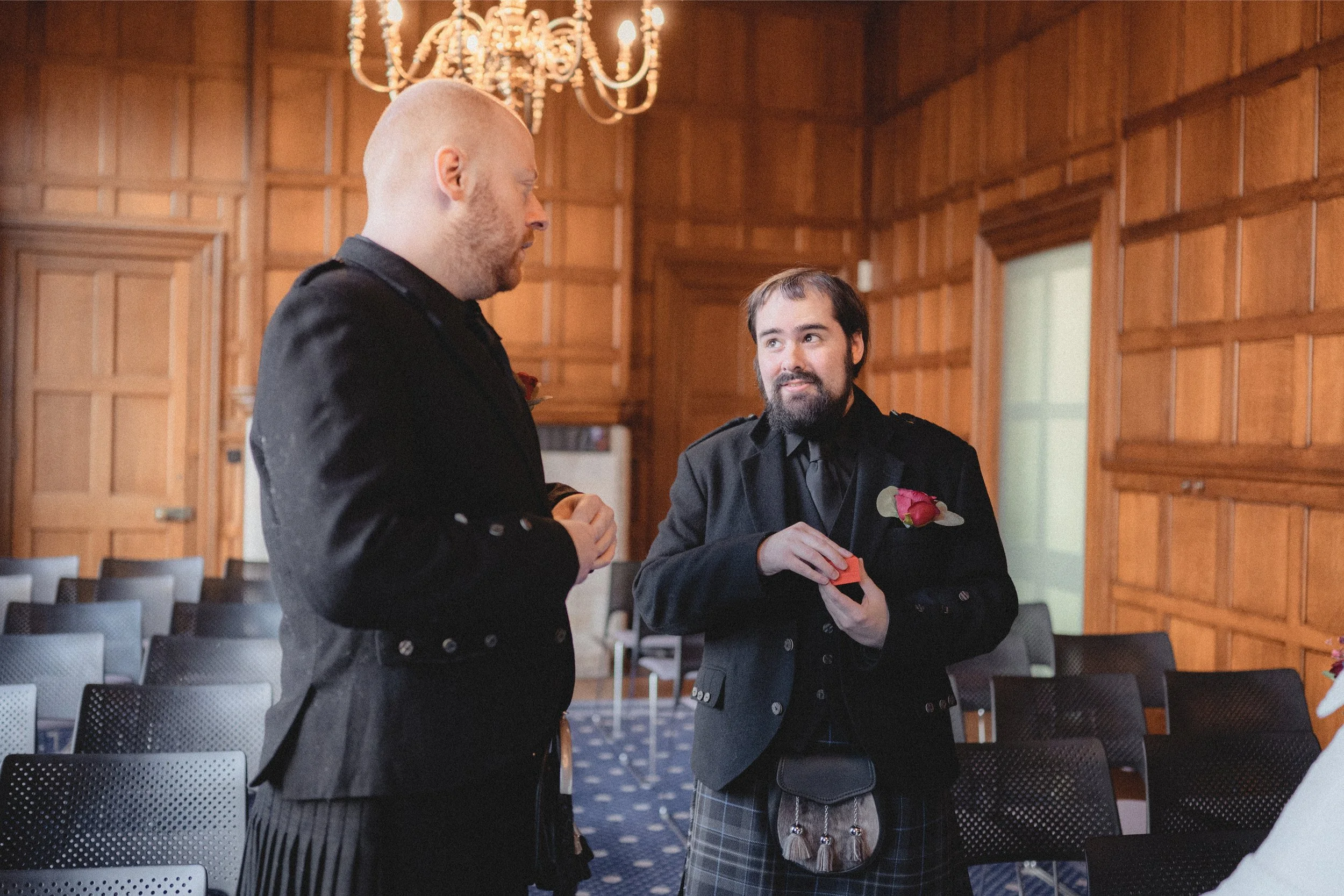 Two men in formal attire engaged in conversation at an indoor event, with wooden-paneled walls, a chandelier, and rows of empty chairs in the background.