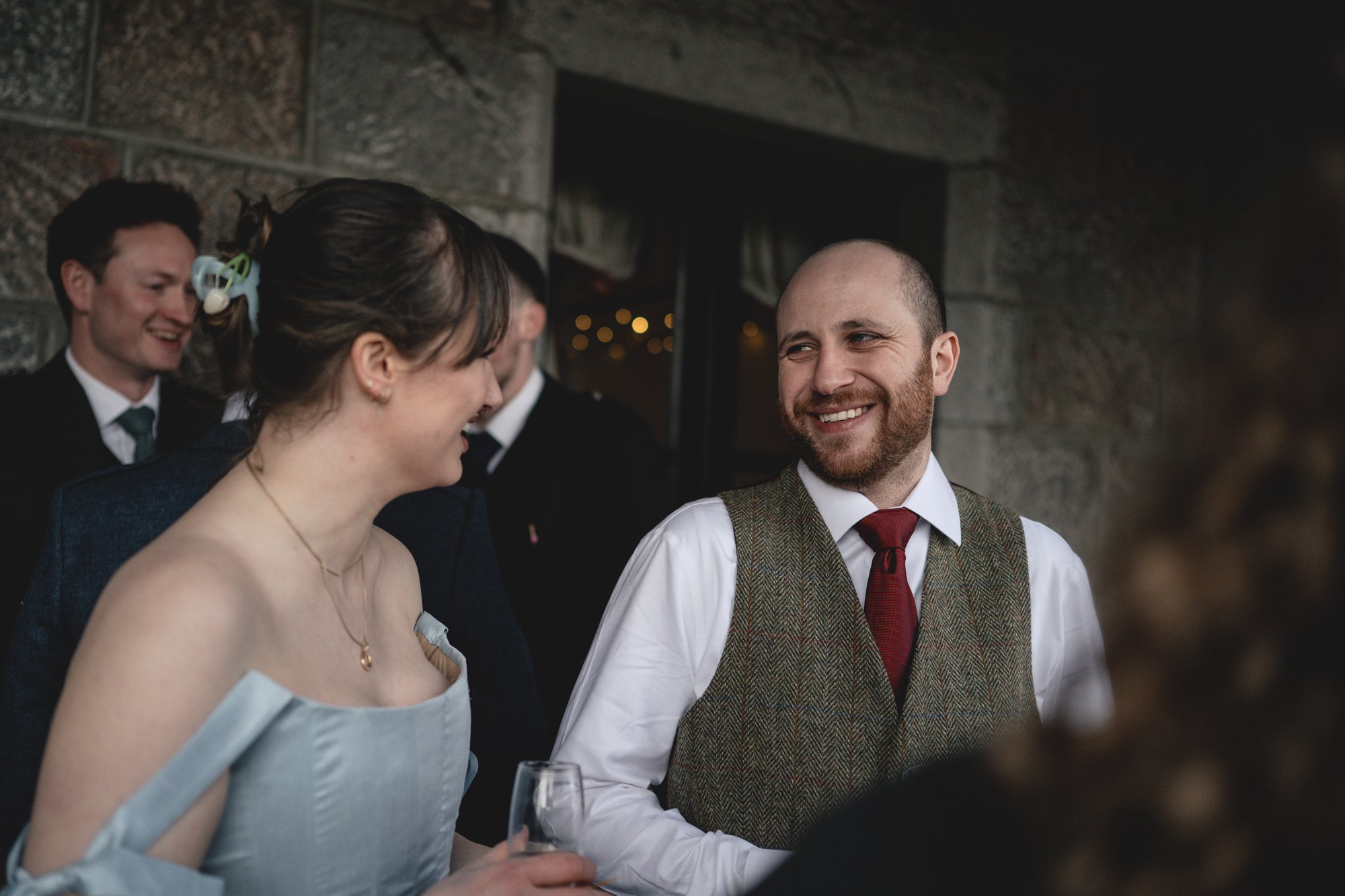 People dressed in formal attire, with some holding glasses, engaged in conversation at an indoor event with stone walls.