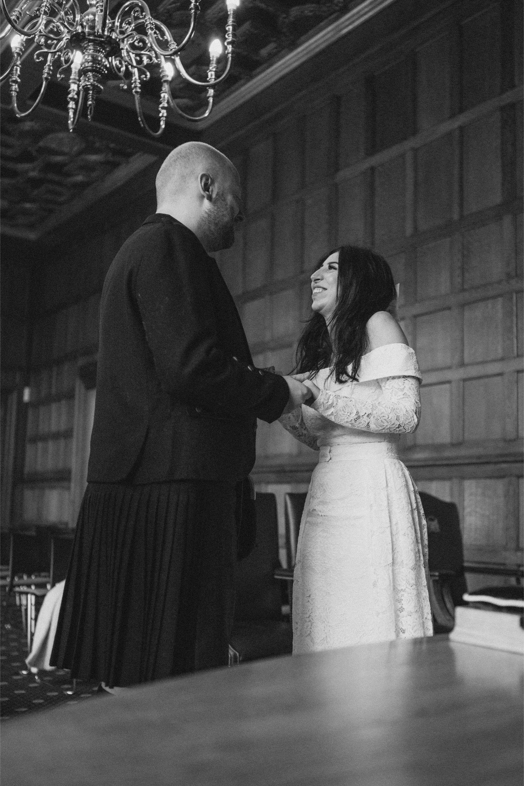 A black and white photo of a couple holding hands and smiling at each other indoors, with a chandelier overhead and wood-paneled walls in the background.
