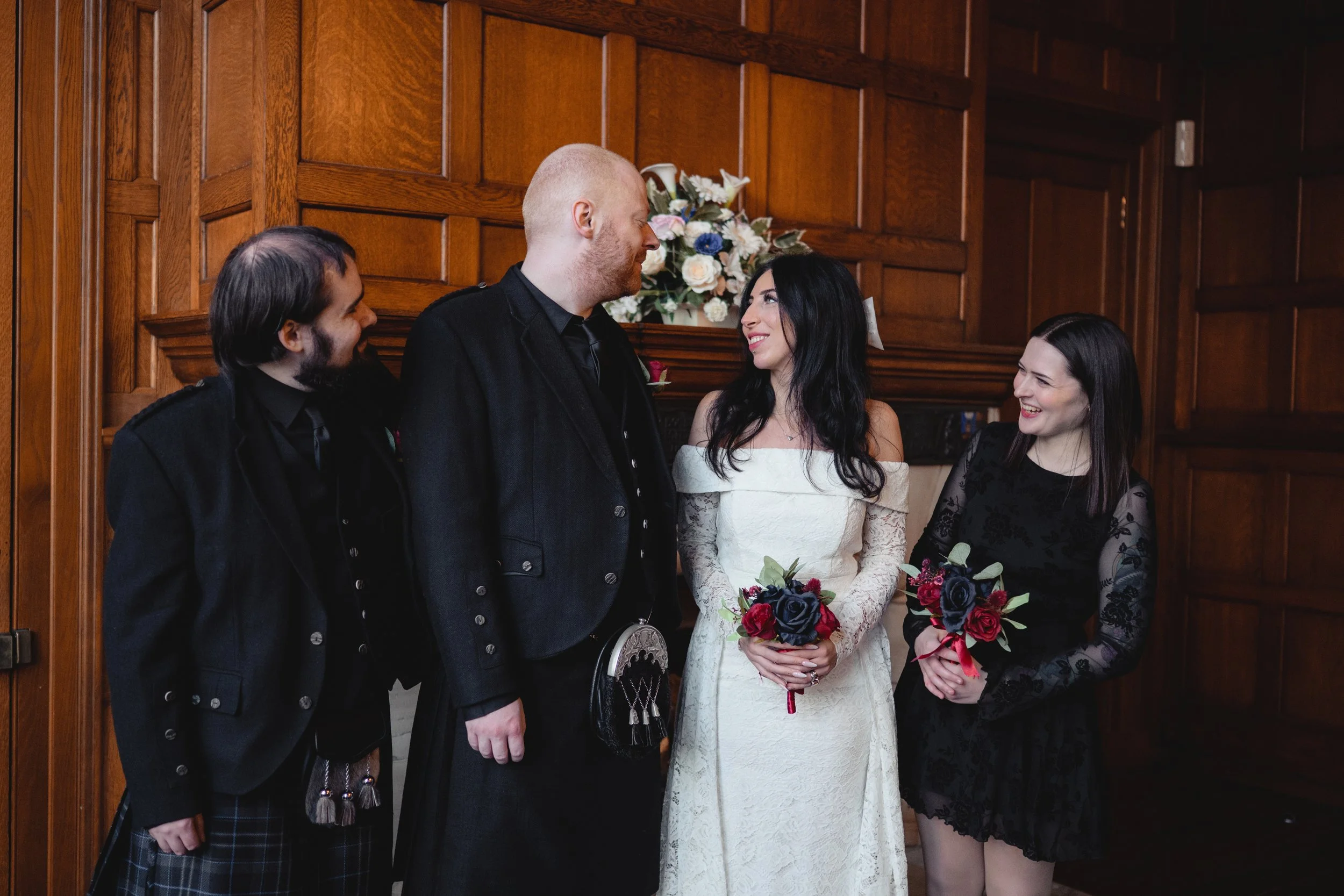 A wedding ceremony with a bride in a white dress holding a bouquet, a groom in a dark suit, and two women in black dresses, all smiling and looking at each other in a wood-paneled room.