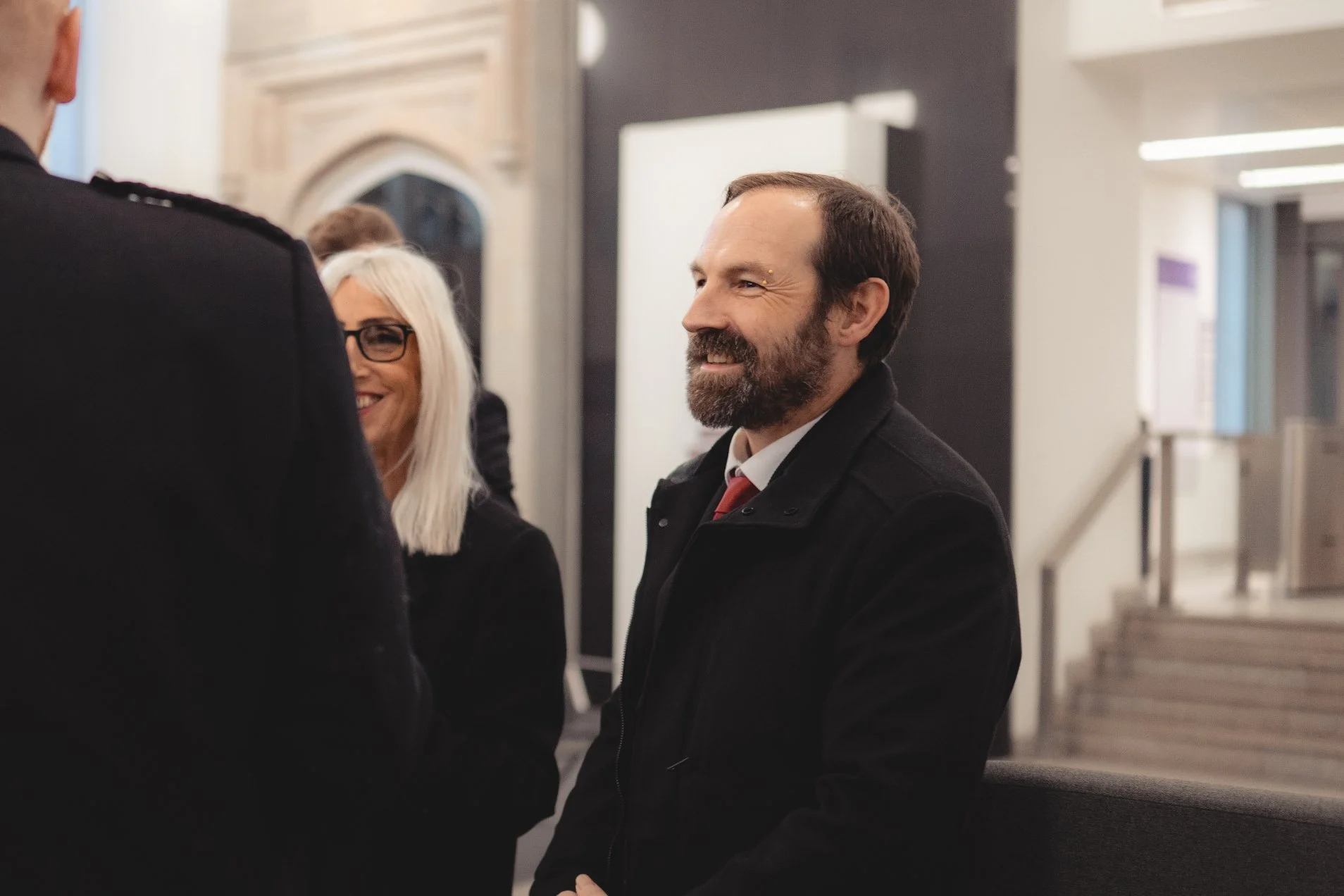 Three people smiling and talking in a public indoor space, including a man with a beard wearing a black coat and a woman with white hair and glasses.