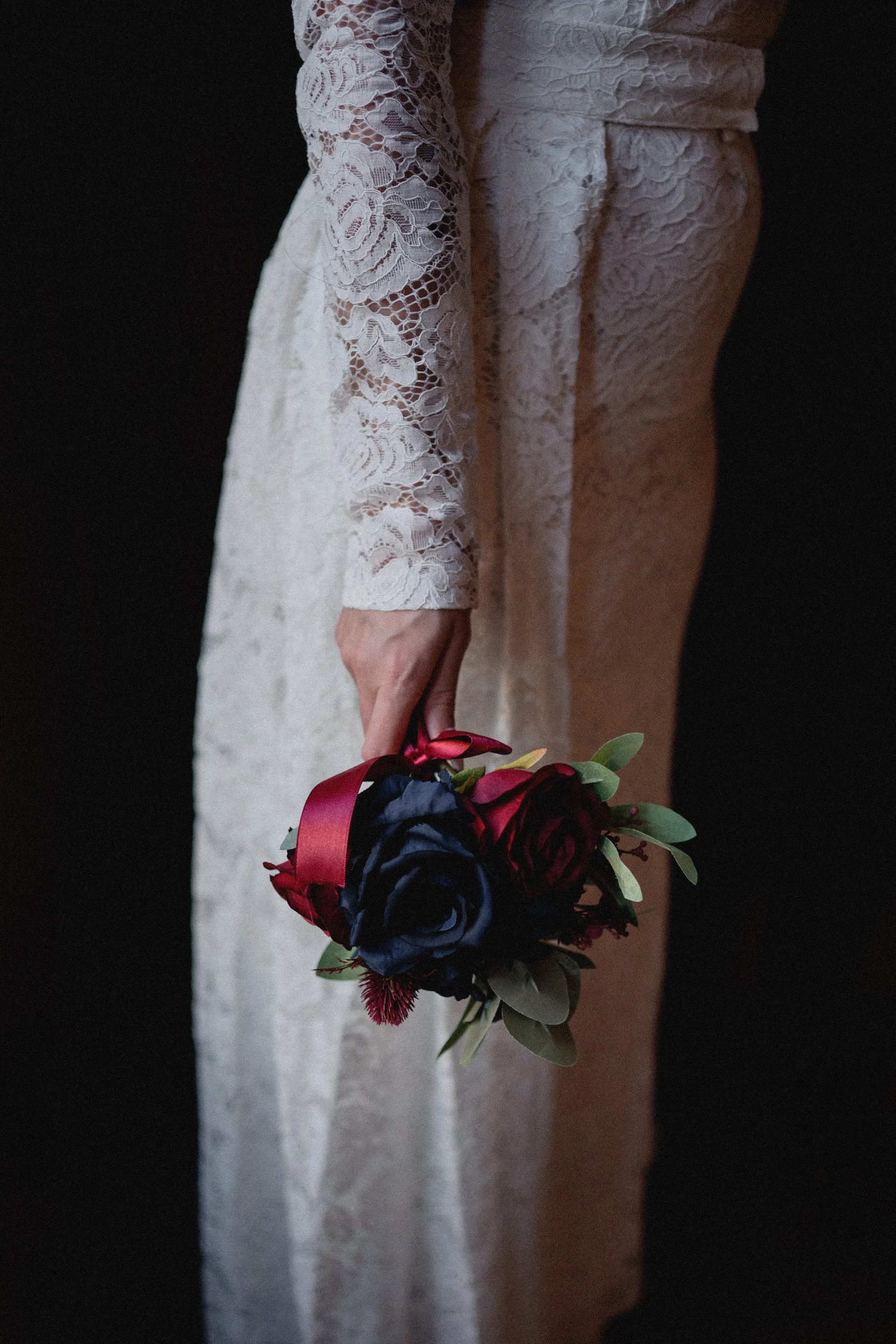 A woman in a white lace dress holding a bouquet of dark red and black roses with green leaves and a red ribbon, against a dark background.