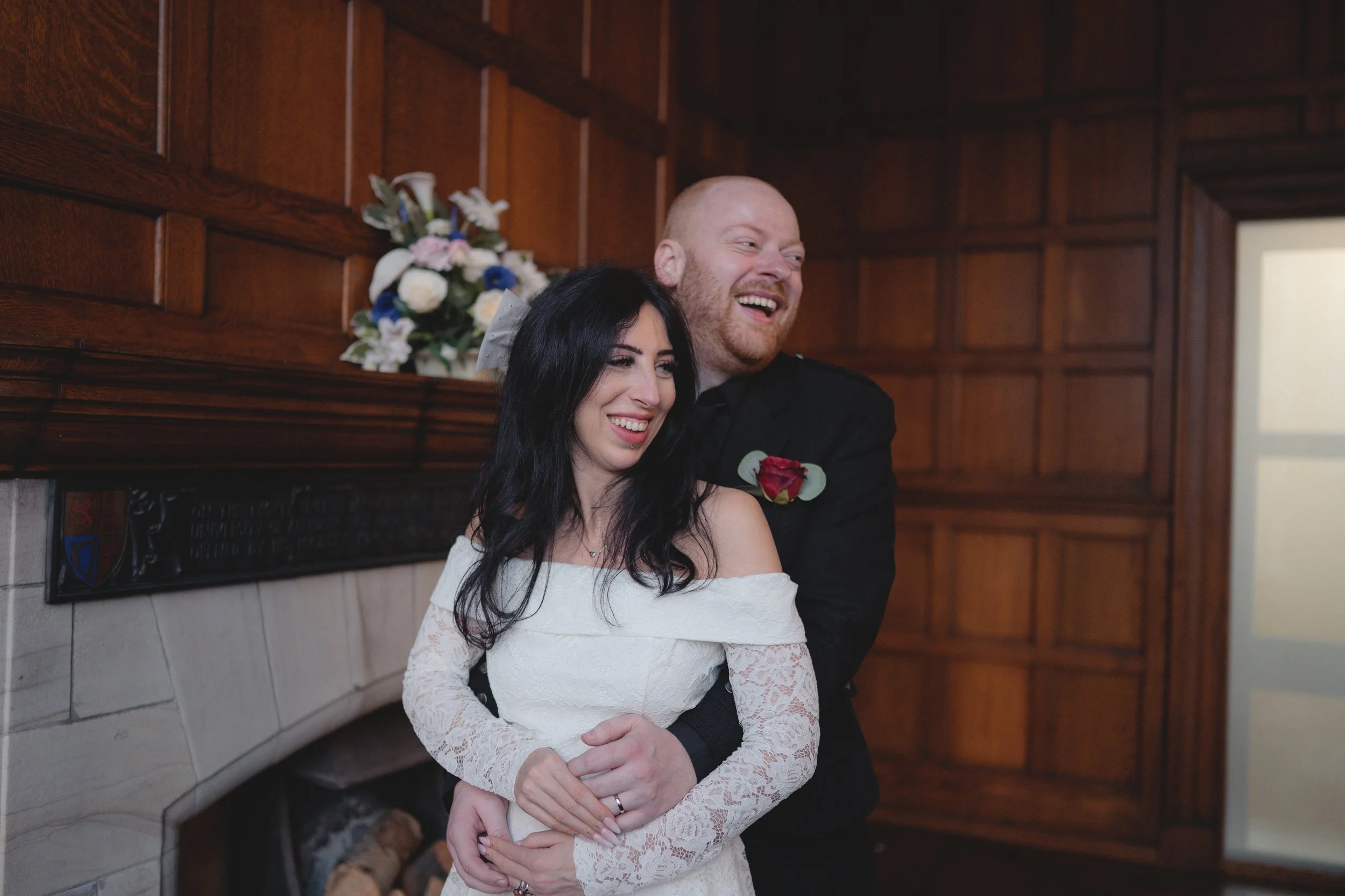 A couple in wedding attire embracing and smiling during their wedding ceremony, with a wooden backdrop and floral arrangement behind them.
