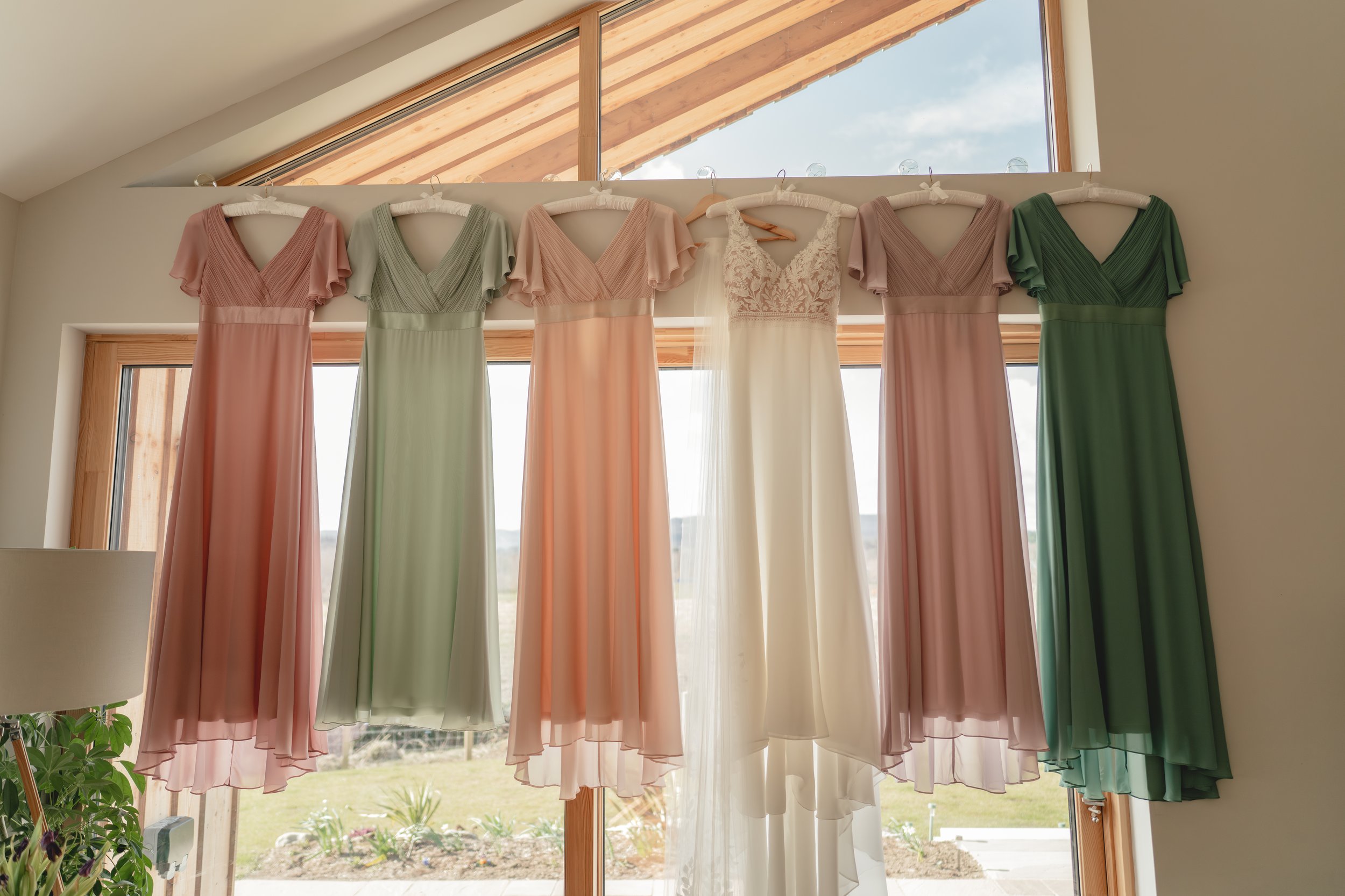 Several pastel-colored dresses hanging on a rack inside a room, with large windows and a glass door showing an outdoor landscape.