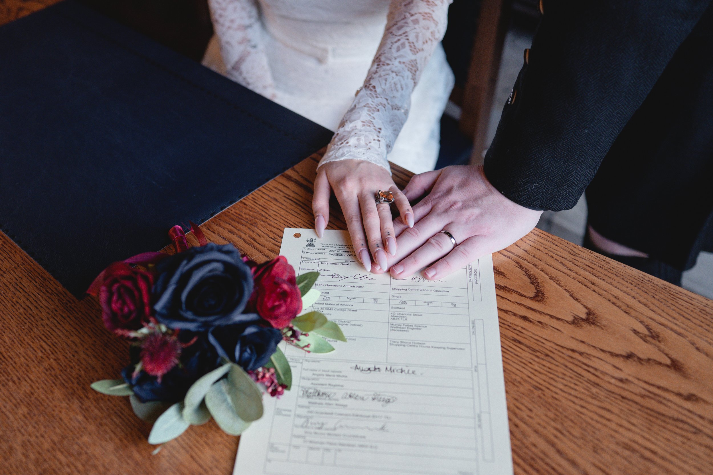 Couple signing marriage license with wedding bouquet on wooden table.