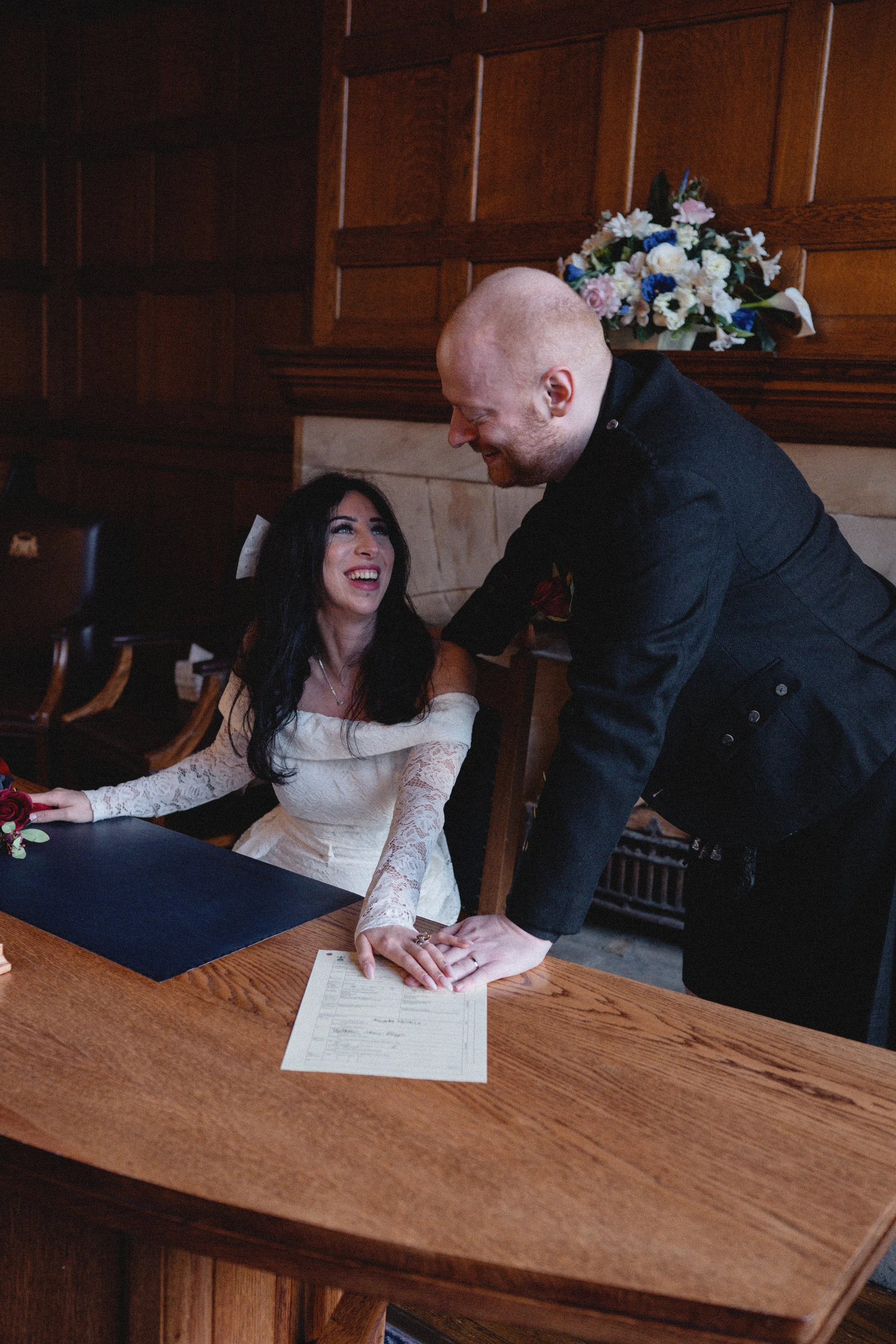 A bride and groom smiling and holding hands over a table during their wedding ceremony. The bride is sitting, wearing a white lace dress, and the groom, standing, is wearing a black jacket. There is a floral arrangement in the background.