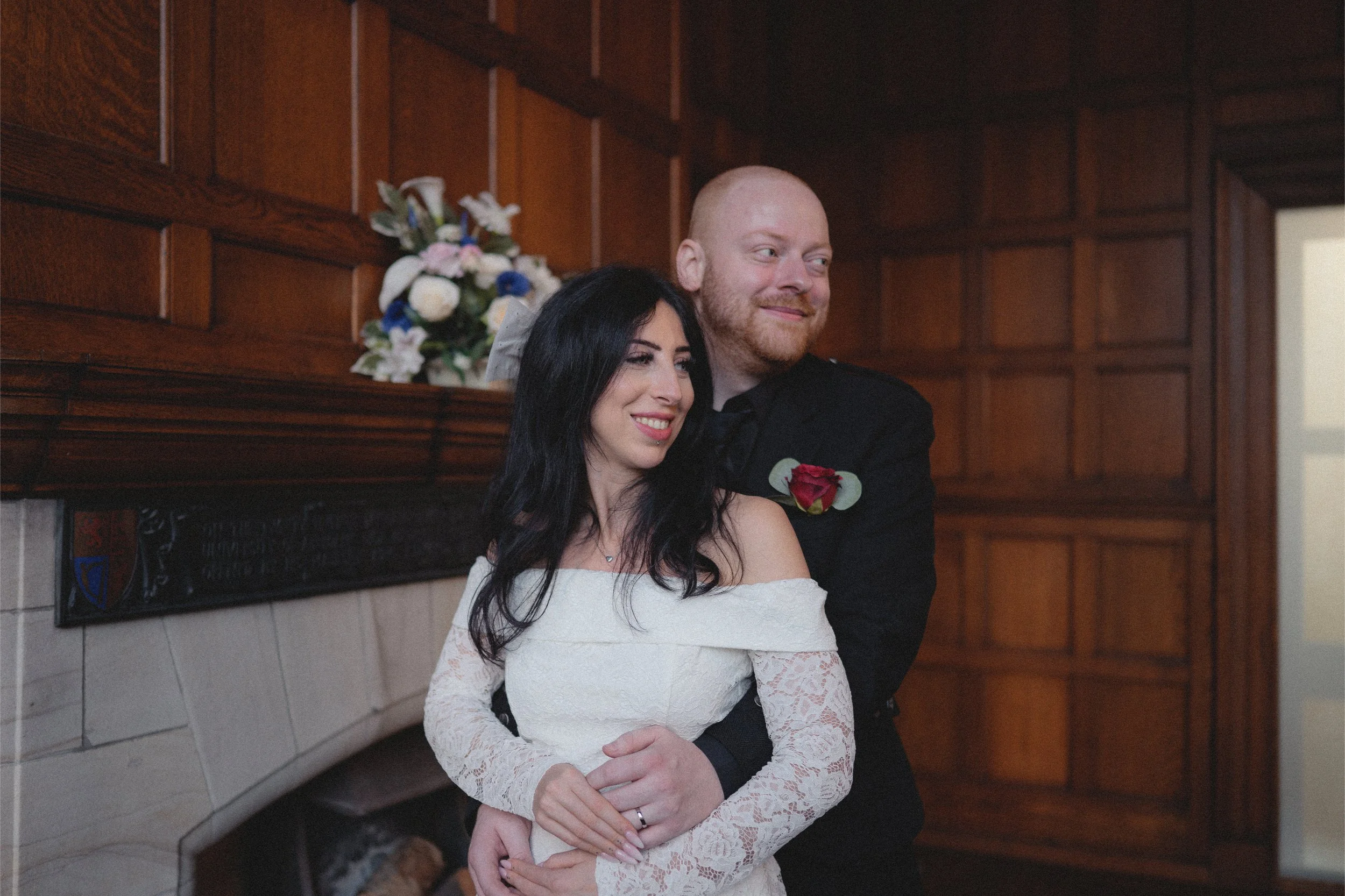 A woman in a white off-shoulder lace wedding dress and a man in a black suit embrace, smiling, inside a wood-paneled room with a floral arrangement on the mantle.