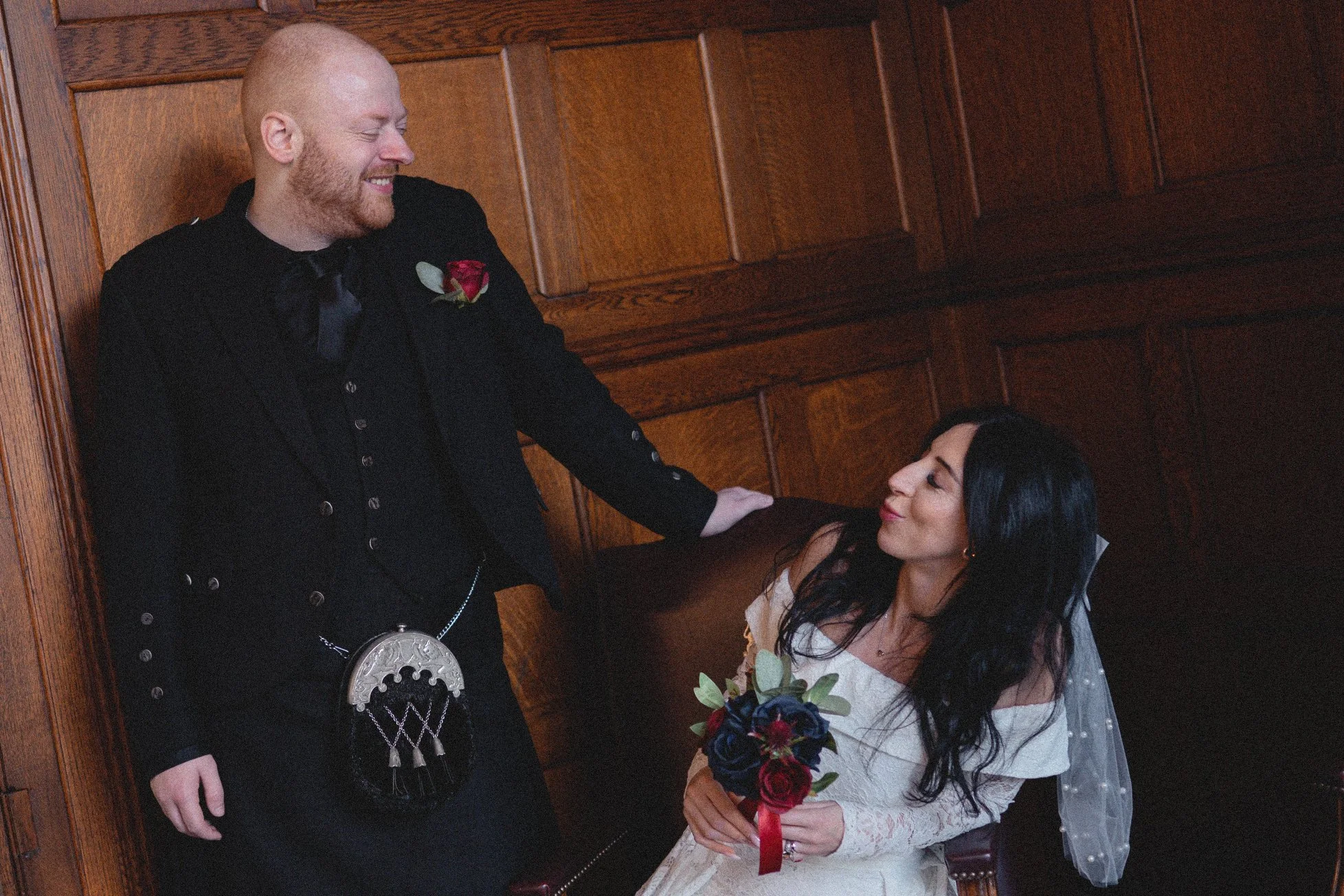 A man in a black suit with a red rose boutonniere and a kilt is smiling and touching a seated woman with long black hair, wearing a white dress and a veil, holding a bouquet of dark roses.