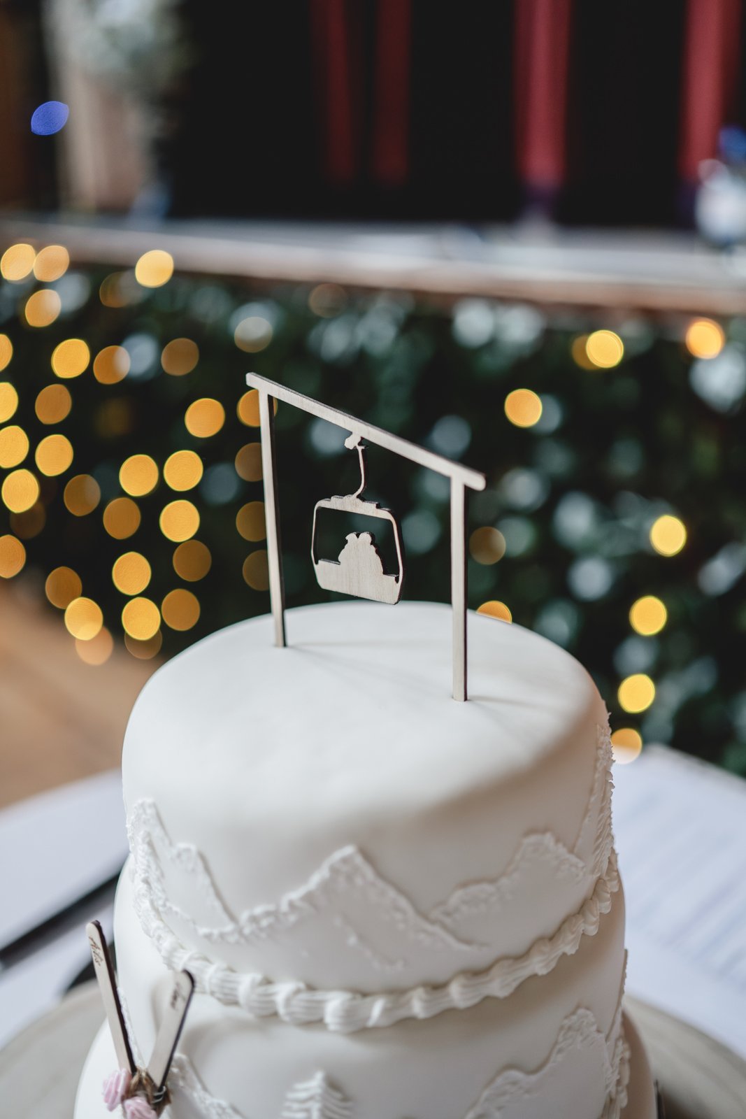 Close-up of a white wedding cake with mountain-like icing design and a metallic cake topper shaped like a ski lift, with Christmas lights in the blurred background.