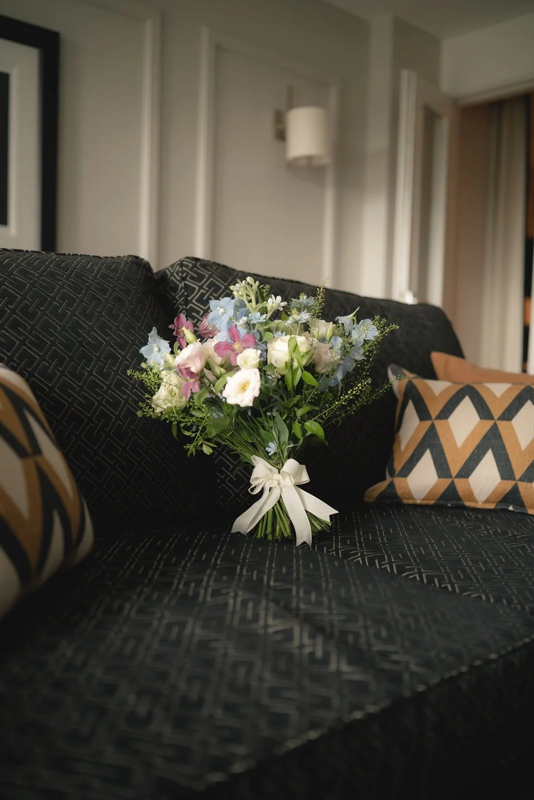 A bouquet of mixed flowers with white, pink, and light blue blooms tied with a white ribbon, resting on a black patterned sofa with geometric-patterned pillows in a living room.