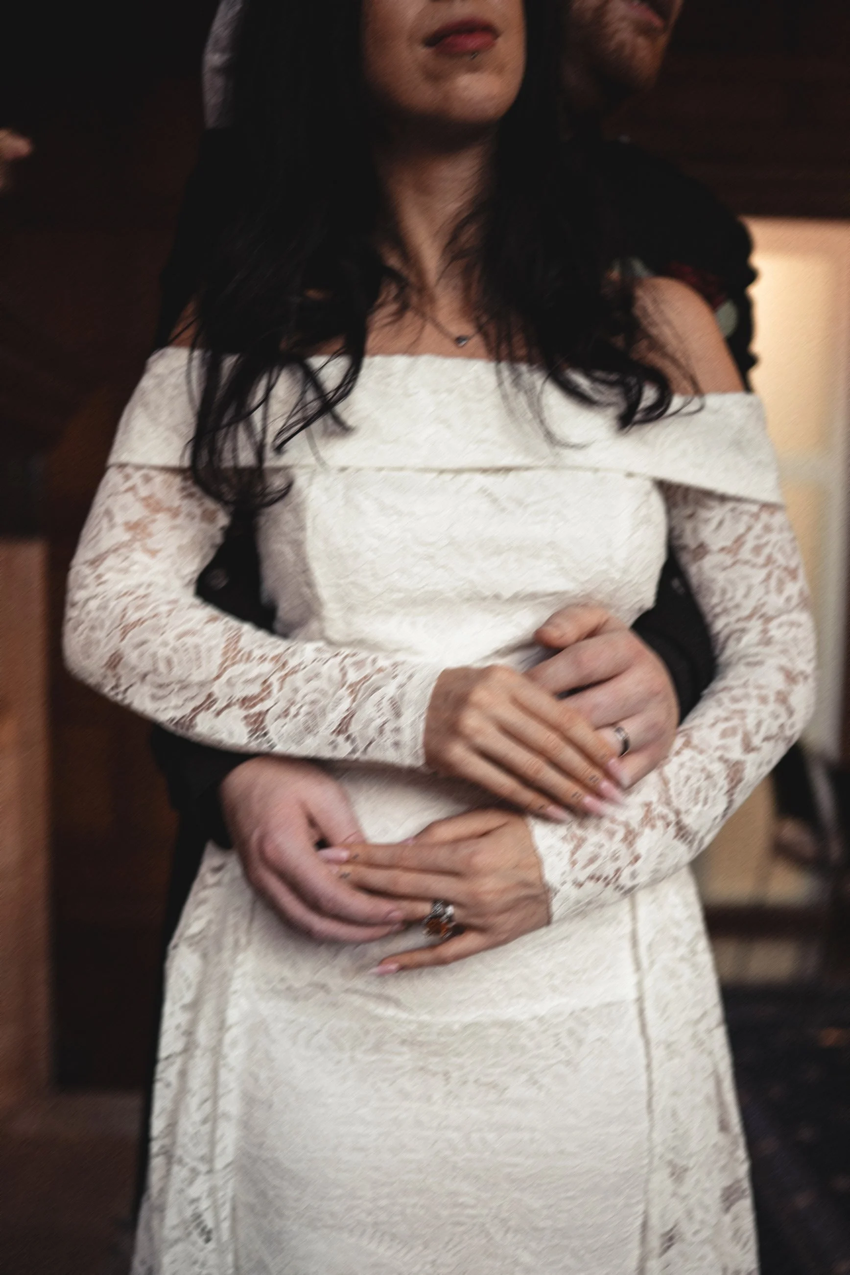 A woman in an off-shoulder lace white dress standing with a man in a black suit behind her, with his arms around her waist.