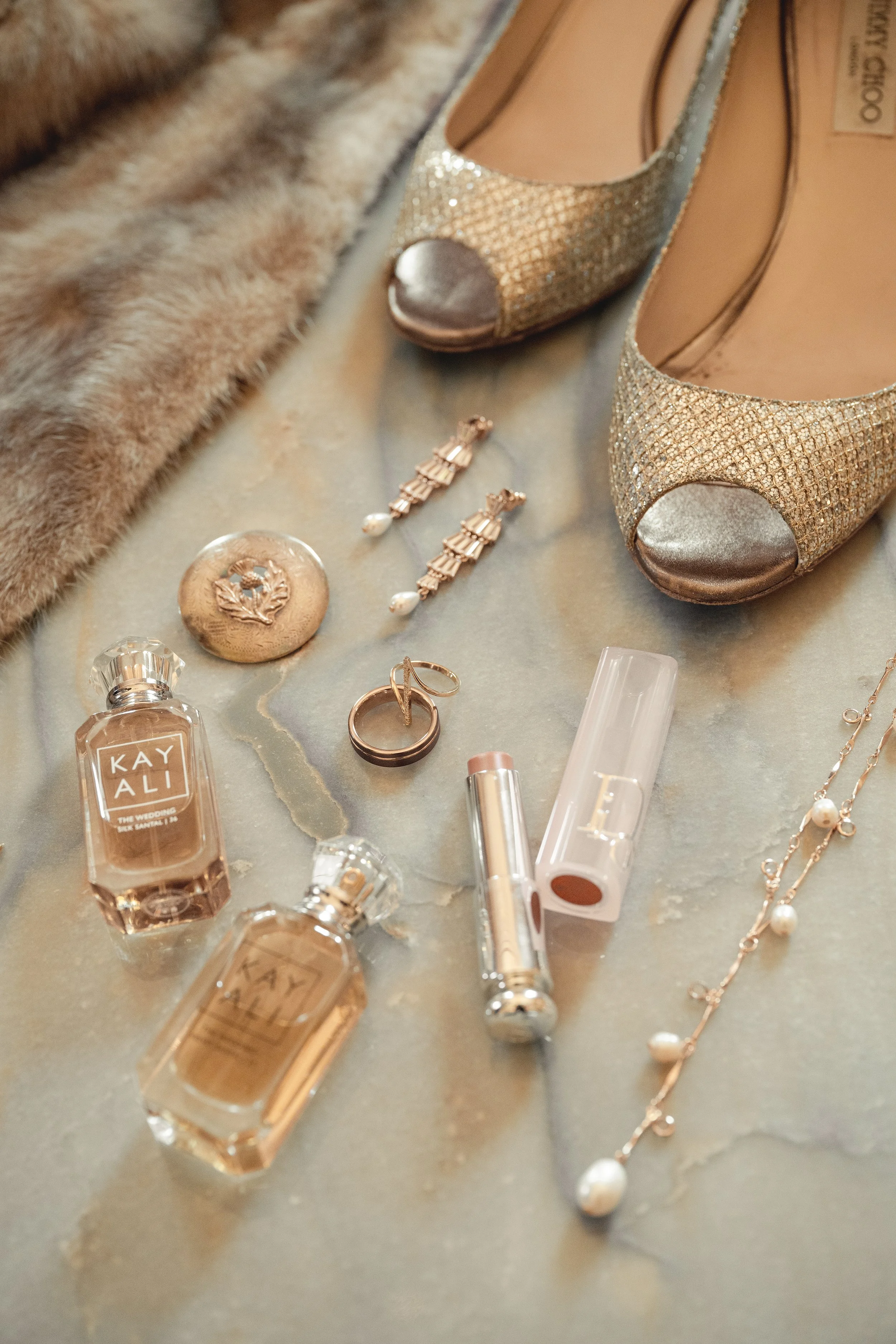 Close-up photograph of various jewelry items and shoes on a surface, including a pair of glittery gold high-heeled shoes, pearl earrings, a pearl necklace, rings, perfume bottles, and other beauty or fashion accessories.