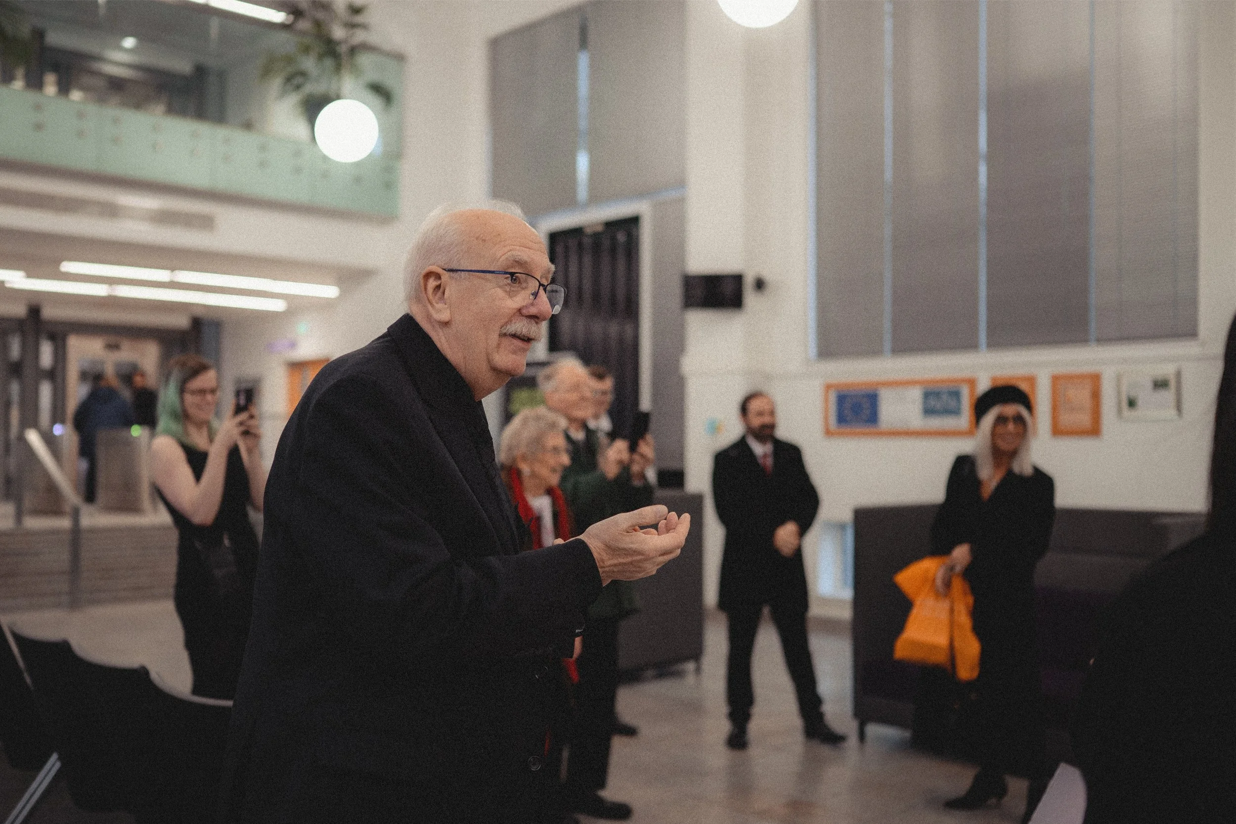 An older man with glasses and a mustache, wearing a black jacket, speaking in a room with several people standing around and taking photos, in a modern indoor space.
