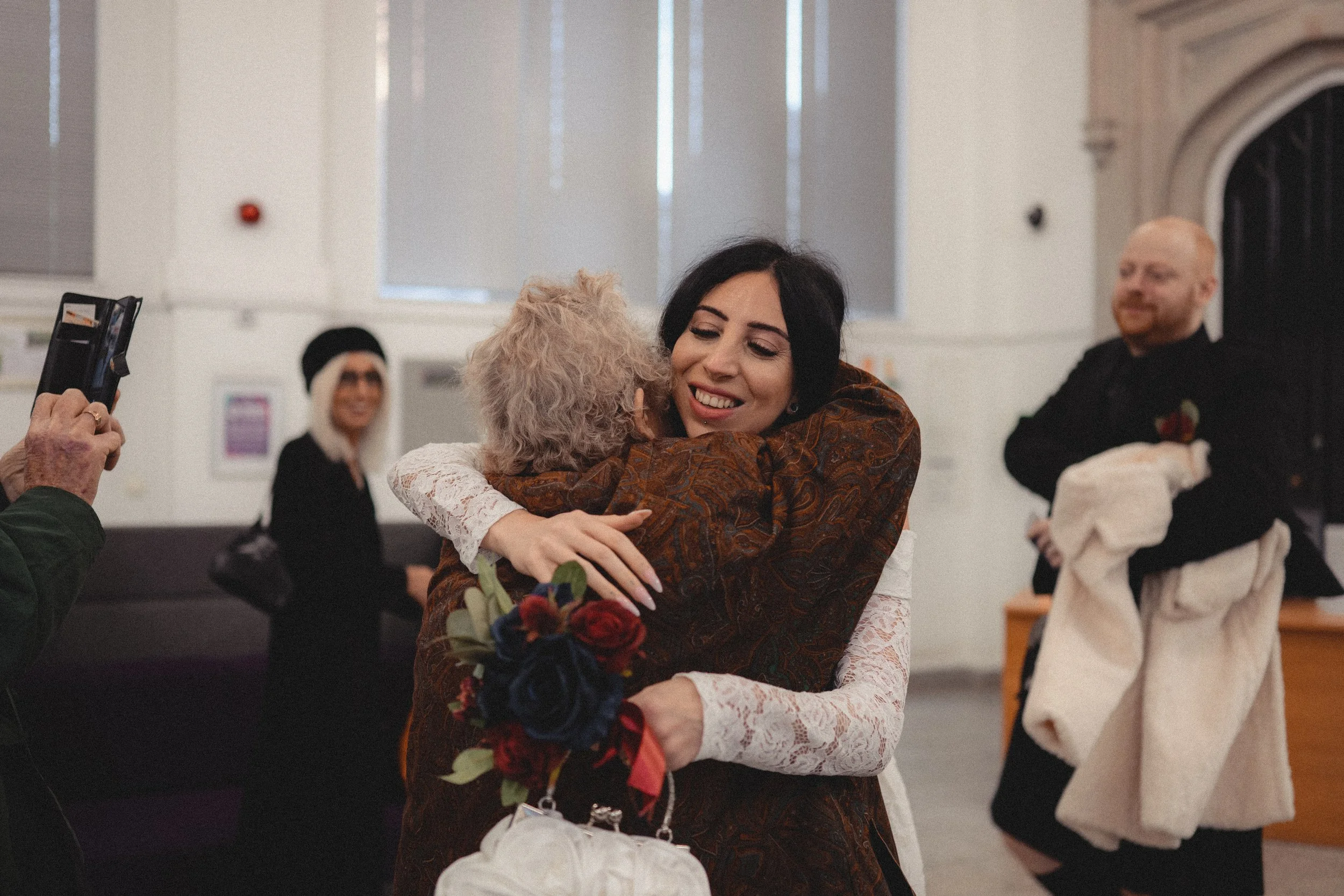 Two women hugging at a welcoming event, with one woman holding a bouquet of flowers. A man on the right holds a coat, and a woman in the background wears a black outfit and hat, smiling. A person on the left captures the moment on a phone.