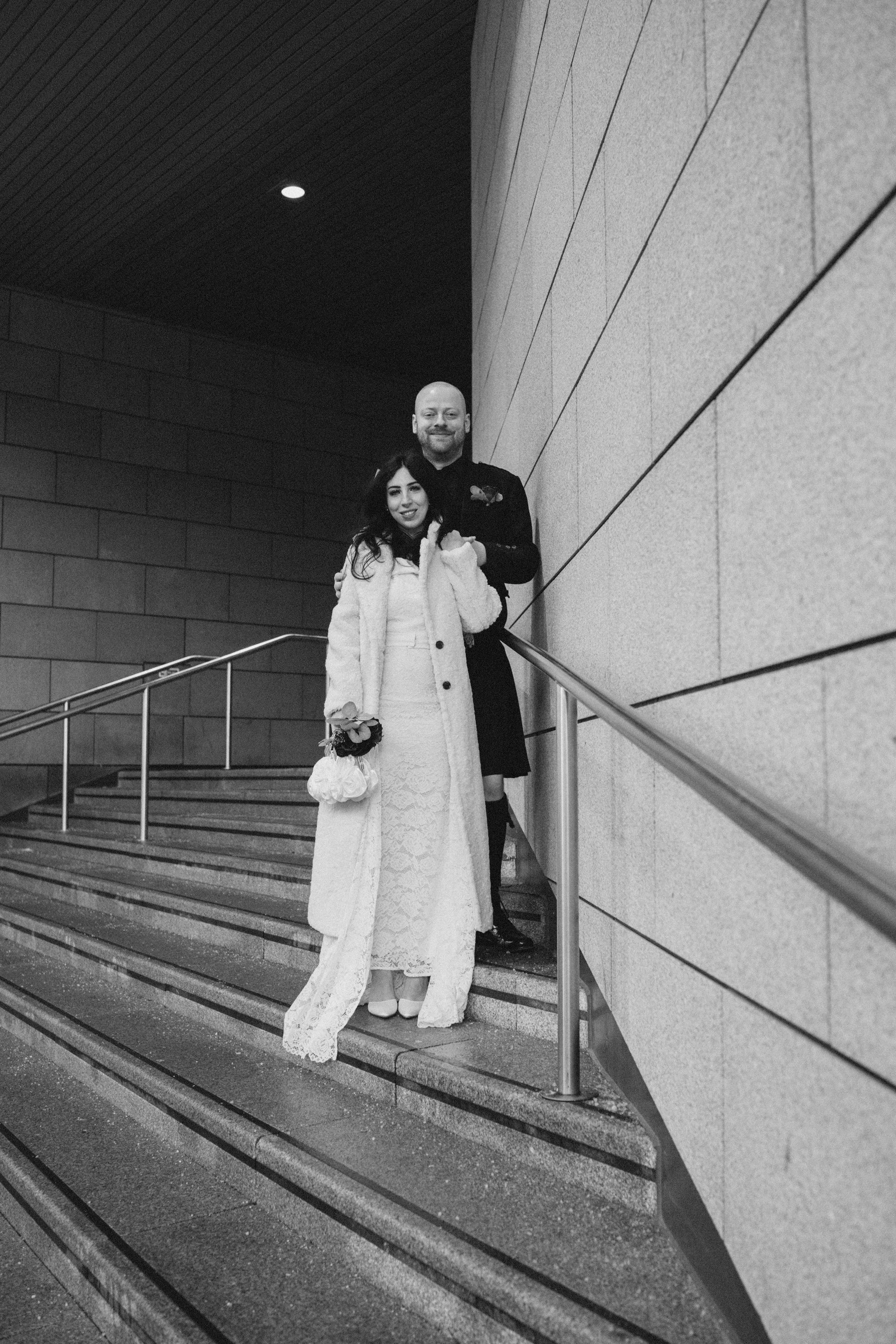 A couple dressed in wedding attire standing on stairs against a modern building wall, smiling and embracing, in black and white.