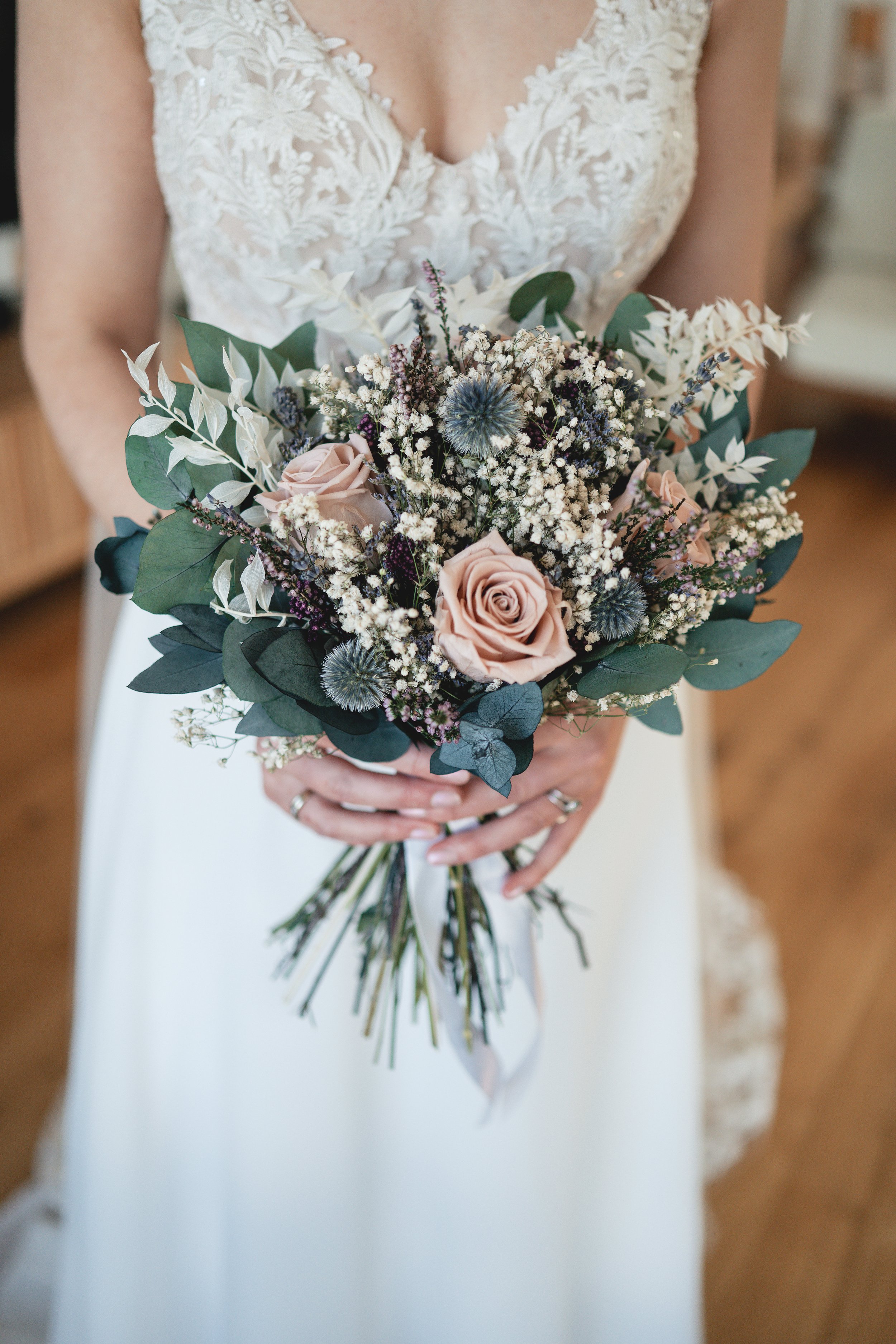 A bride holding a bouquet of various flowers, including pink roses, white baby's breath, and greenery, in front of her dress with lace detailing.