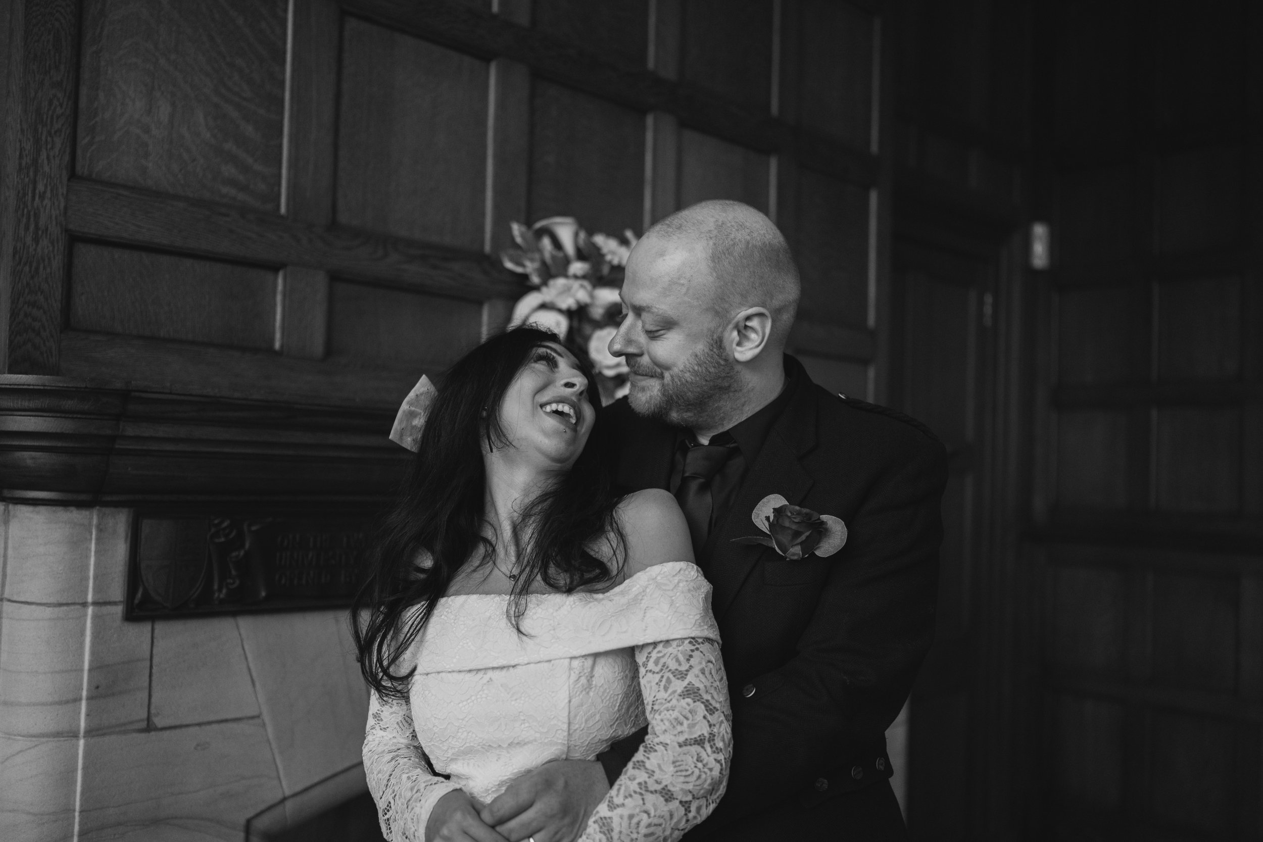 Black and white photo of a woman and man on their wedding day, smiling and embracing in an indoor setting.