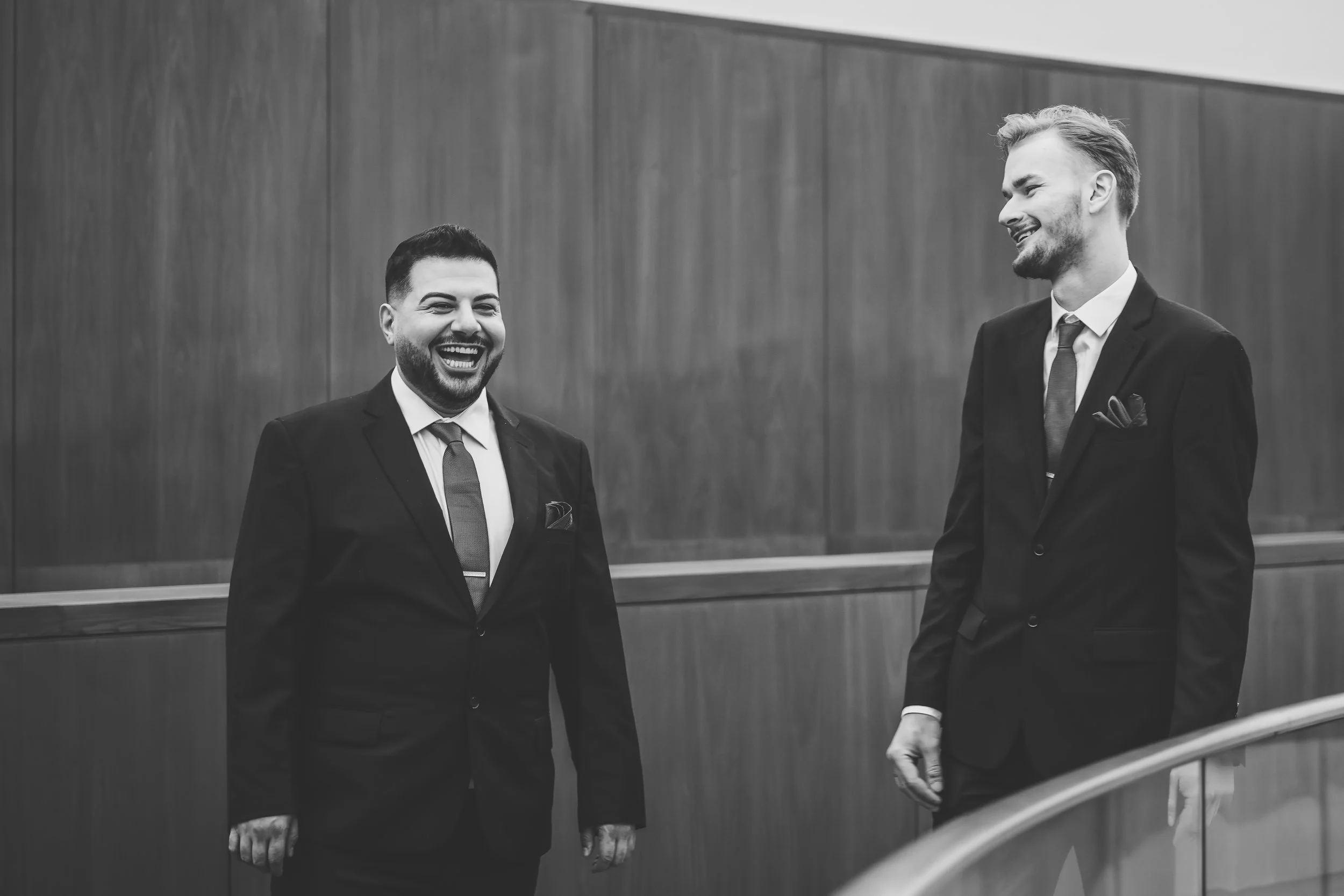Two men in business suits are laughing and smiling at each other in a professional setting with wooden wall paneling.
