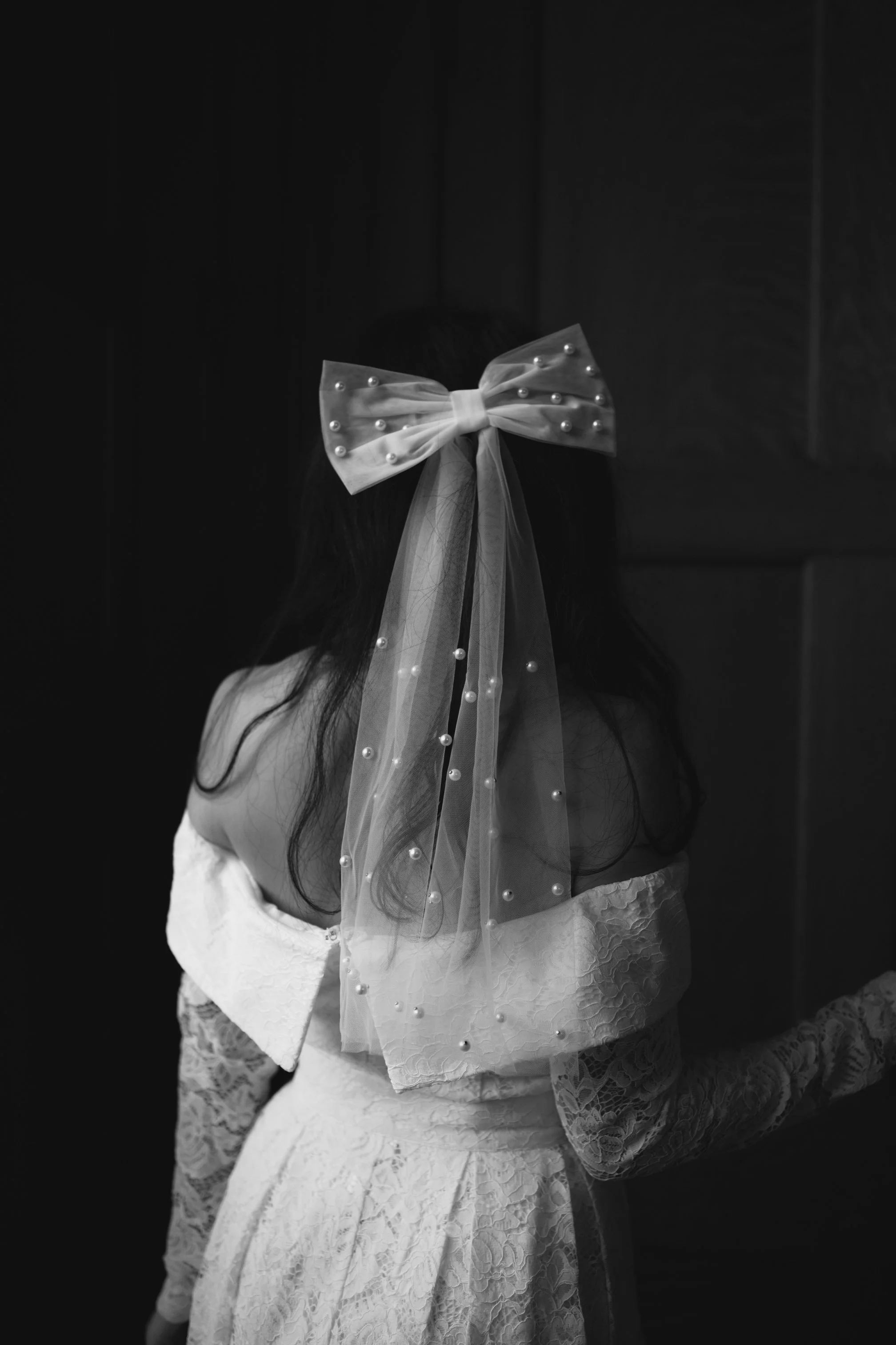 A woman seen from behind wearing a lace dress with a sheer veil decorated with pearls and a large bow on her head.