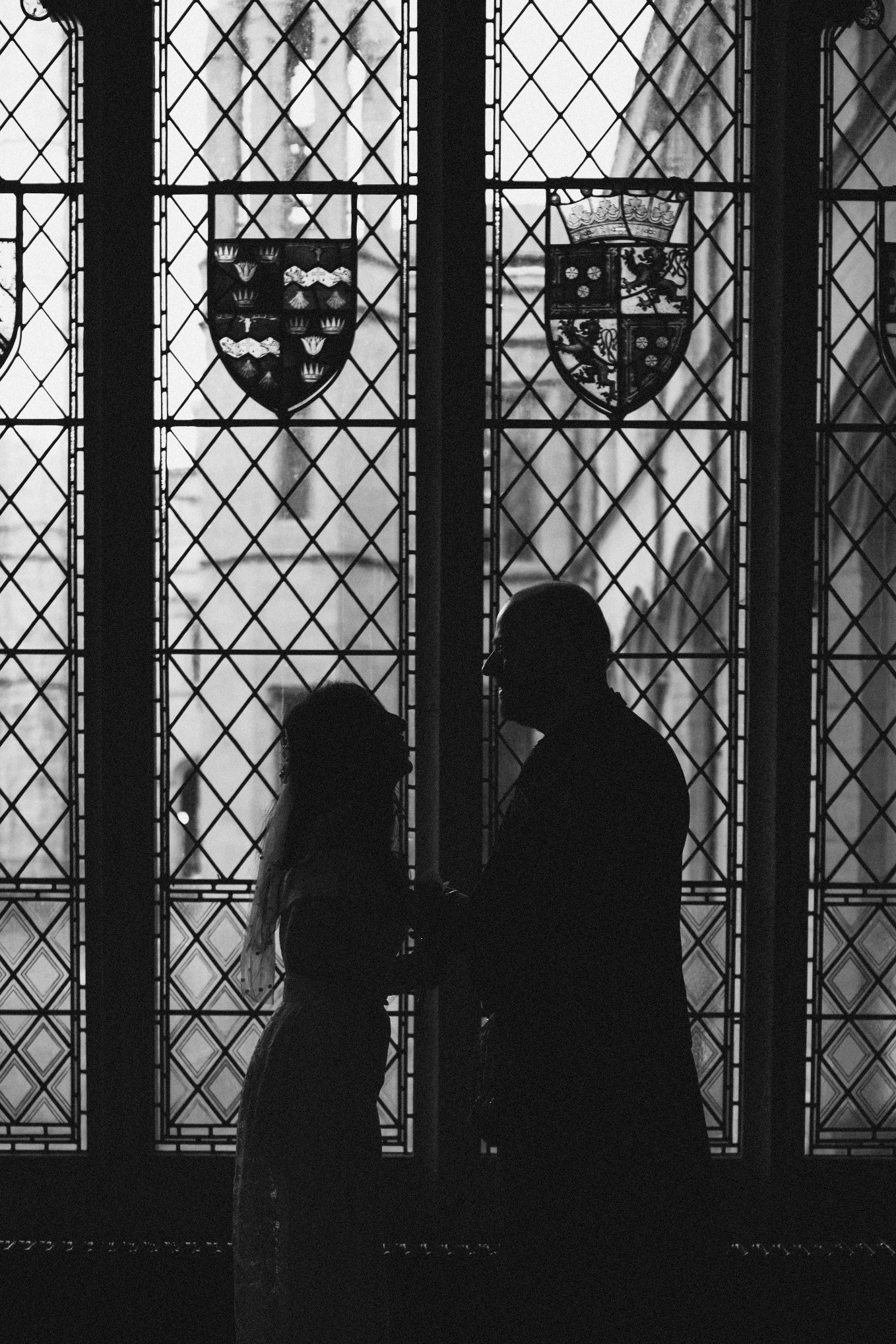 Silhouettes of a man and woman holding hands in front of stained glass windows with family crests, inside a building.