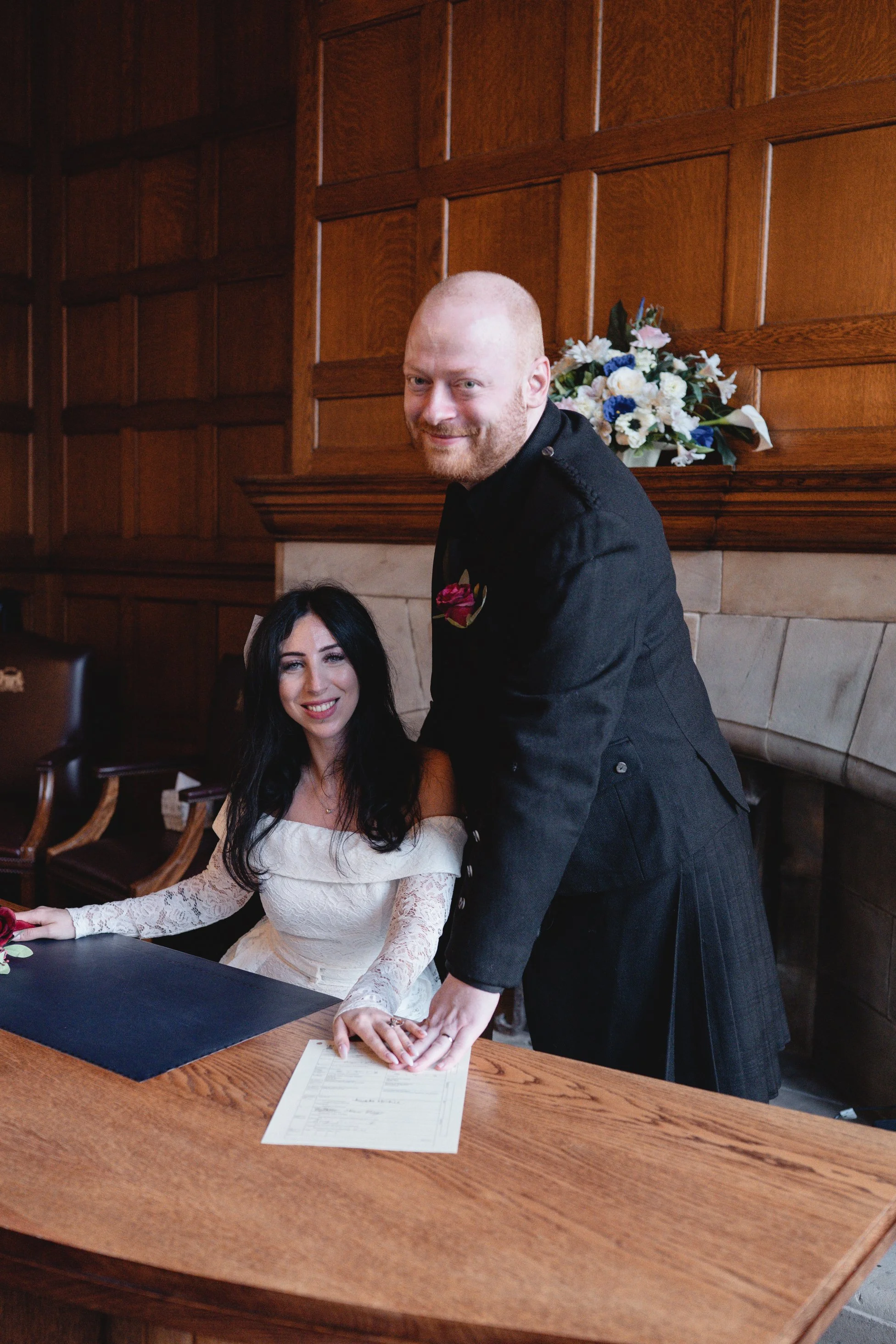 A woman in a white off-shoulder lace wedding dress sitting at a wooden table, smiling, as a man in a black kilt and jacket signs a document. There are floral arrangements and wood-paneled walls in the background.