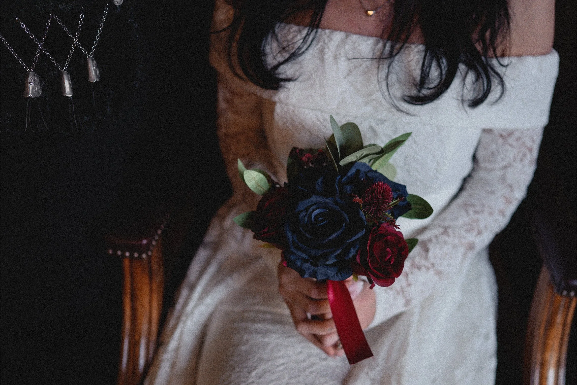 A bride holding a dark blue and red bouquet of flowers while seated. She is wearing a white lace wedding dress with an off-the-shoulder design. The photo is taken from above, focusing on her bouquet and dress.