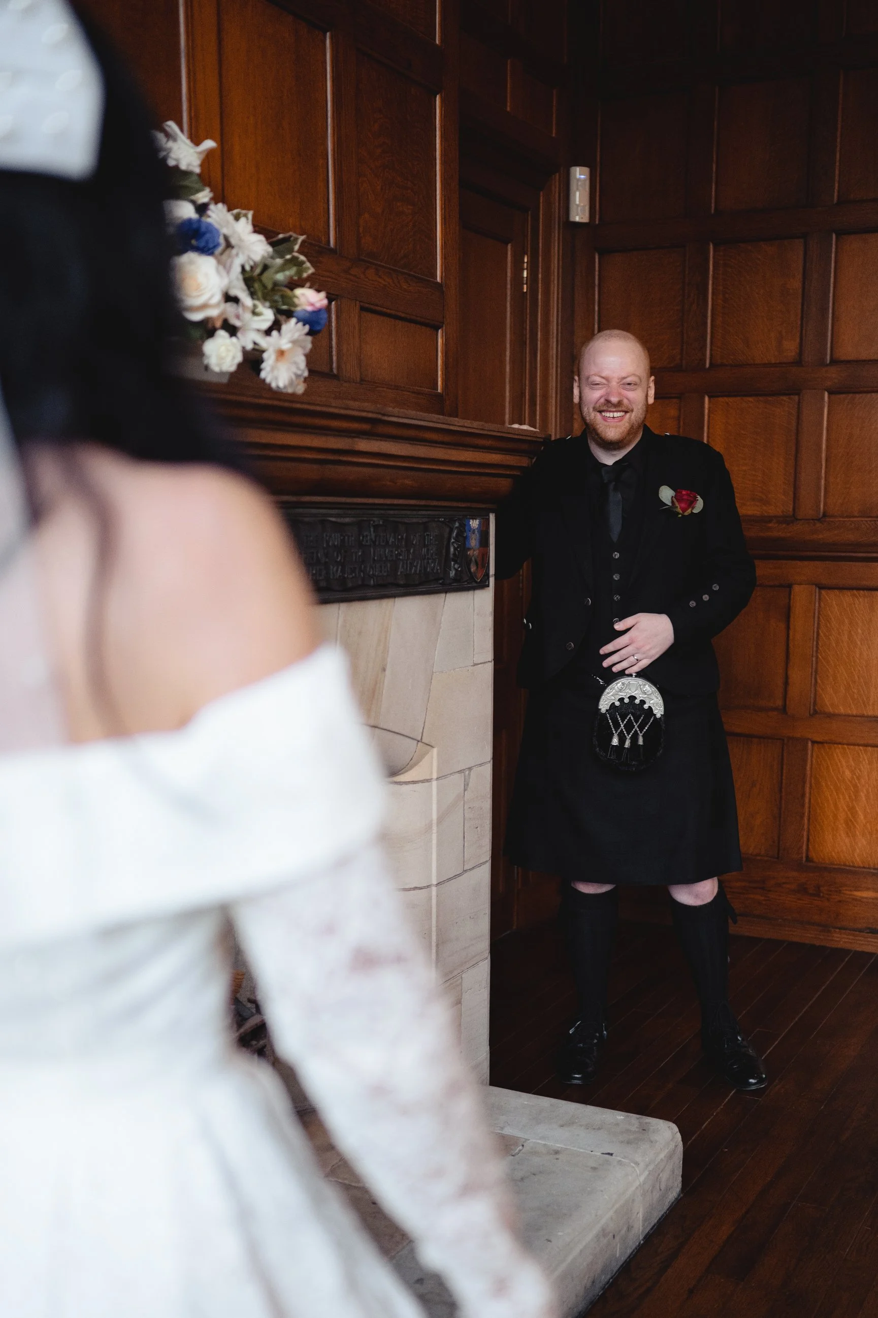 A man in traditional Scottish attire, including a kilt, standing next to a fireplace with a wooden-paneled wall behind him, smiling and looking at a woman in a white dress.