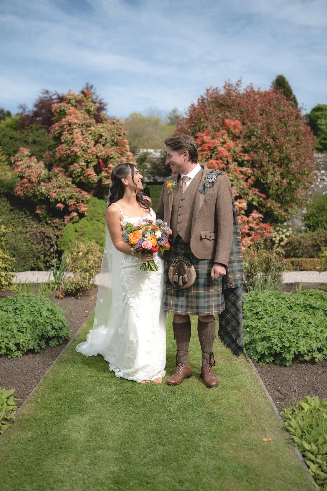 A newlywed couple in wedding attire standing in a garden with colorful flowering trees and green plants, smiling at each other.