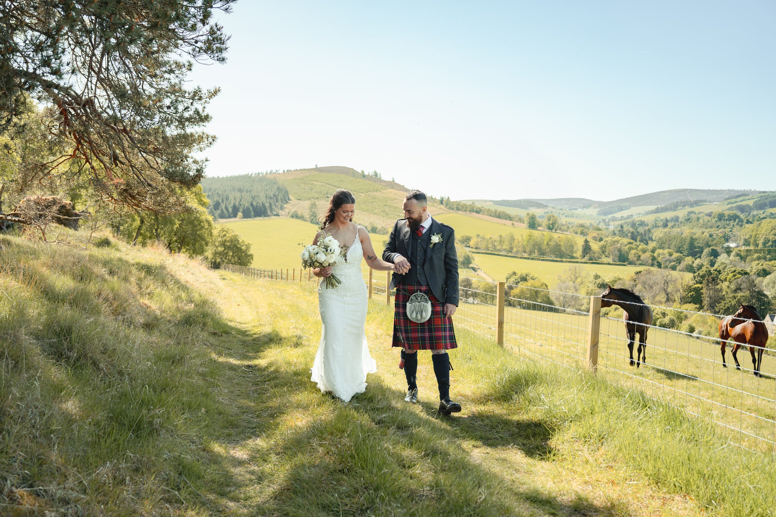 A bride and groom walking hand in hand through a scenic green field with trees and hills in the background. The groom is dressed in traditional Scottish attire, including a kilt, and the bride is holding a bouquet of white flowers, both smiling.