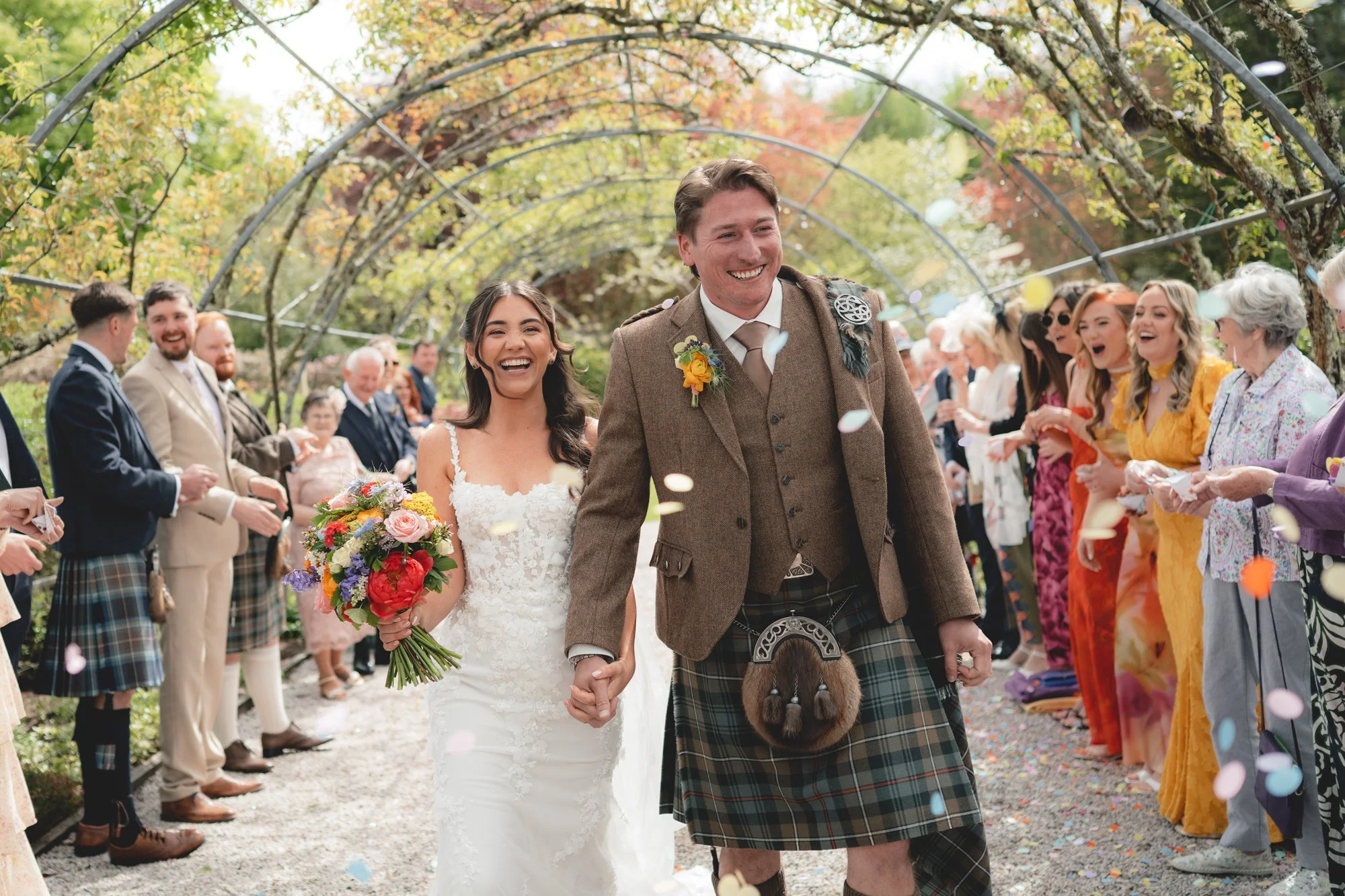 A newly married couple is walking down the aisle at an outdoor wedding, surrounded by friends and family, under a canopy of flowering trees. The bride is holding a colorful bouquet and smiling, while the groom is dressed in traditional Scottish attir