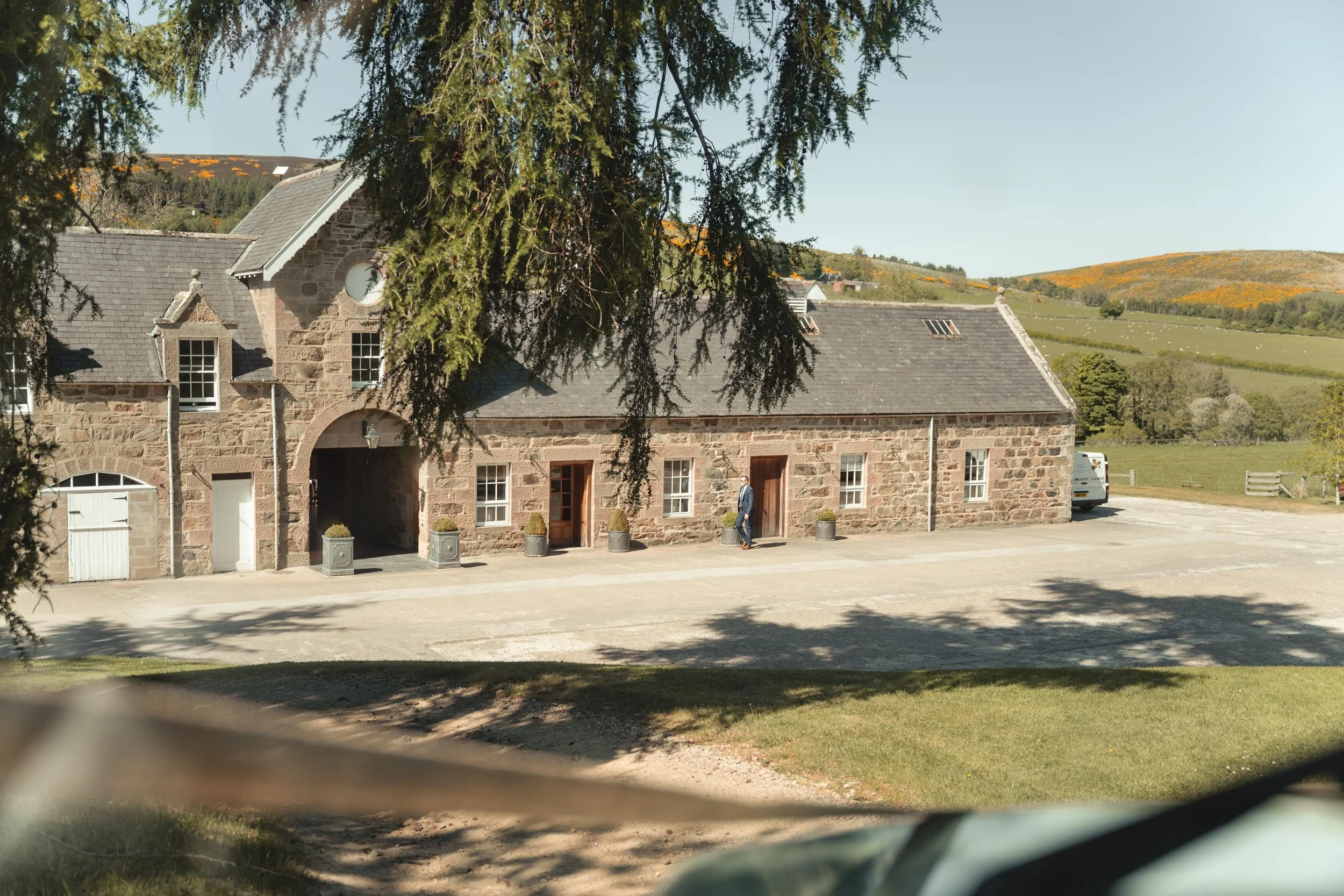 A large stone building with multiple windows and doors, surrounded by a driveway and parking area. In the background, rolling hills with green and yellow foliage are visible under a clear blue sky. A person is standing near one of the doors.