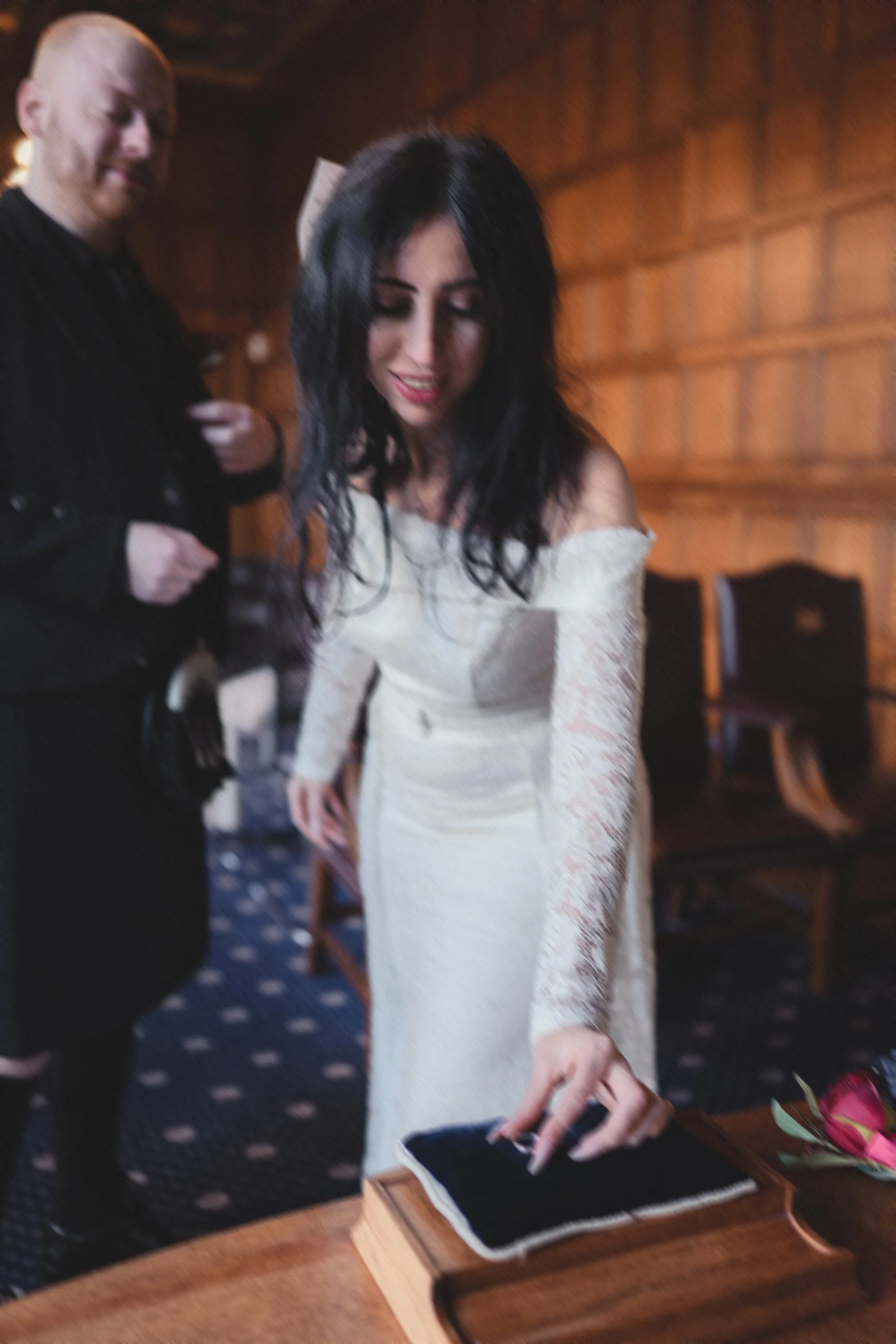 A woman dressed in a white off-the-shoulder lace dress is pointing at a black book on a wooden table, with a woman with short hair in black standing beside her in a wood-paneled room.