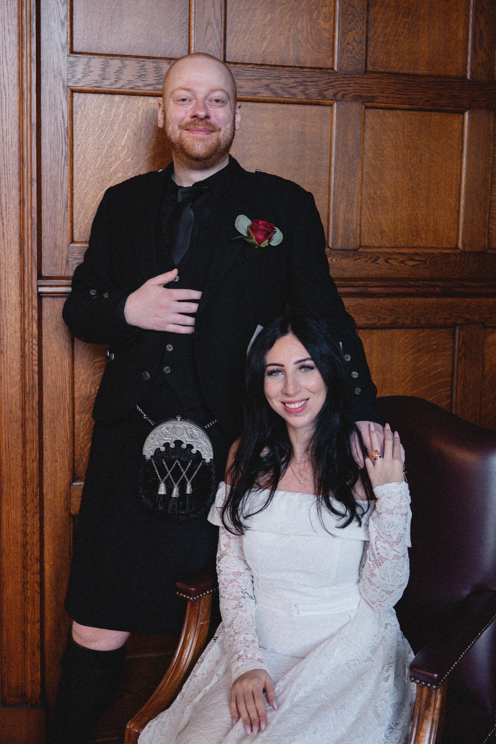 A man in a black suit with a red rose boutonniere standing beside a seated woman in a white lace wedding dress, both smiling inside a wood-paneled room.