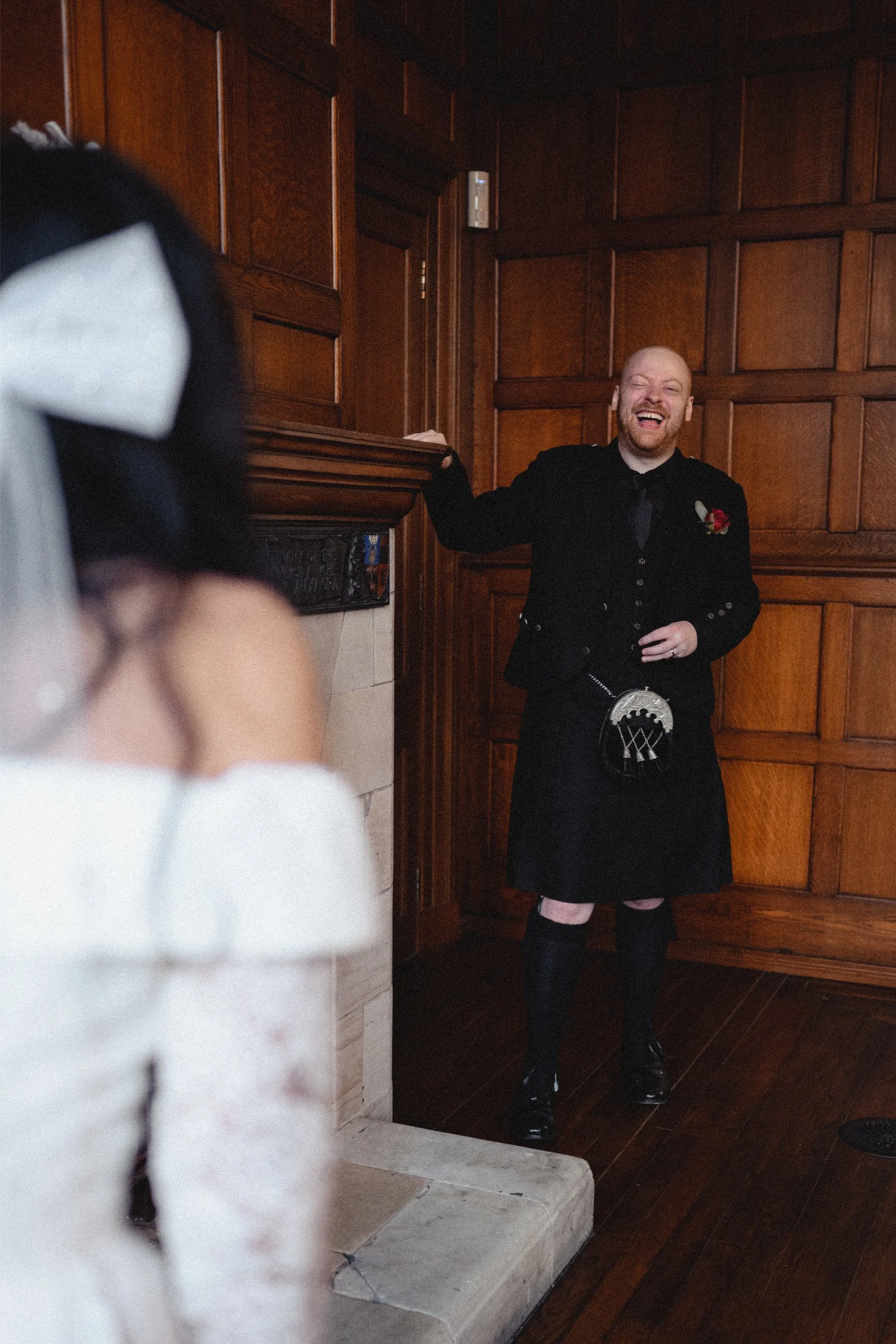 A man dressed in traditional Scottish attire, including a kilt, standing in a wood-paneled room, smiling and laughing while looking at a woman in a wedding dress, who is partially visible in the foreground.