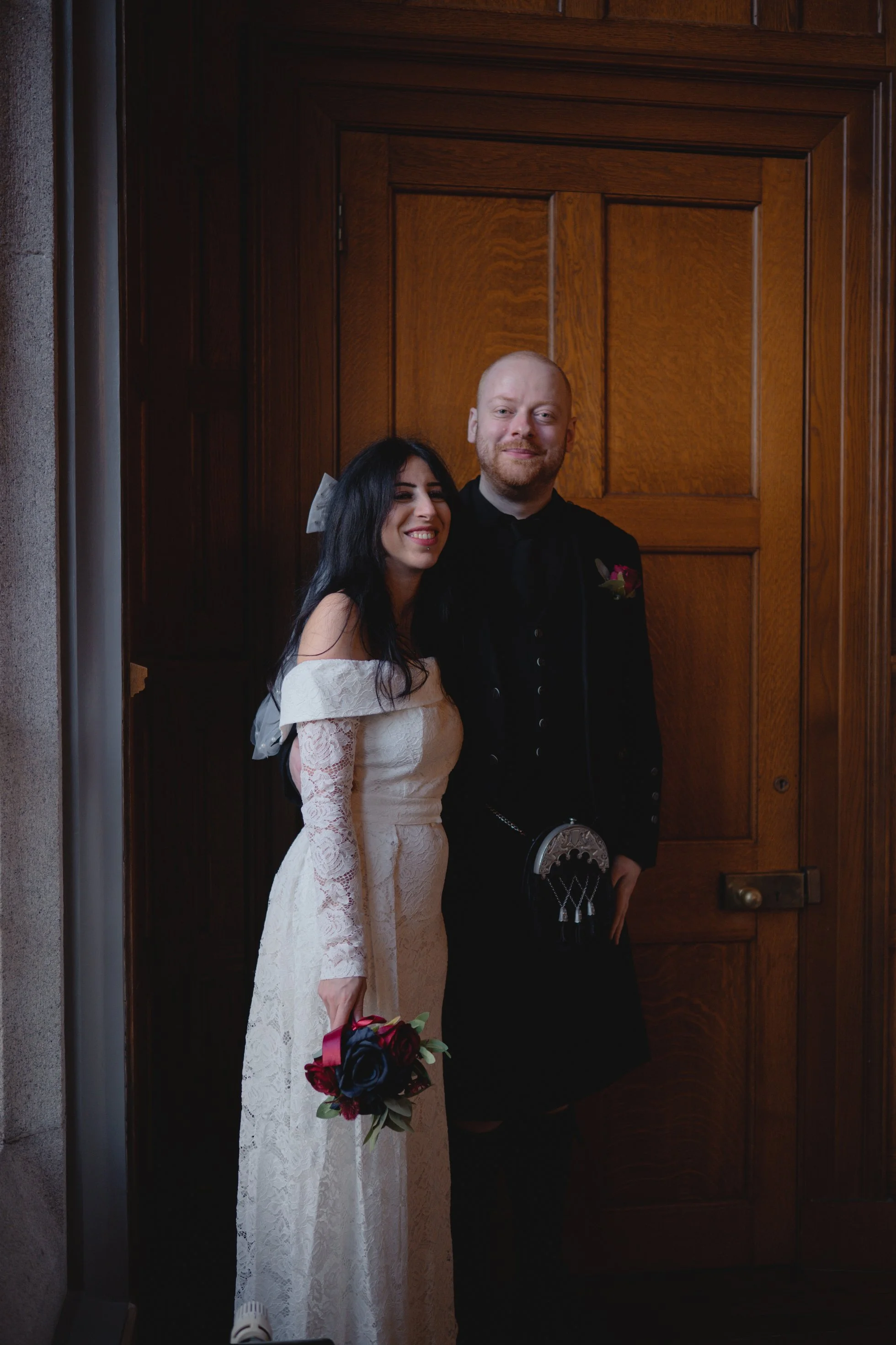 A bride and groom standing together in front of a wooden door, smiling for a photo. The bride wears a white lace gown and holds a bouquet, while the groom is dressed in traditional Scottish attire, including a kilt.