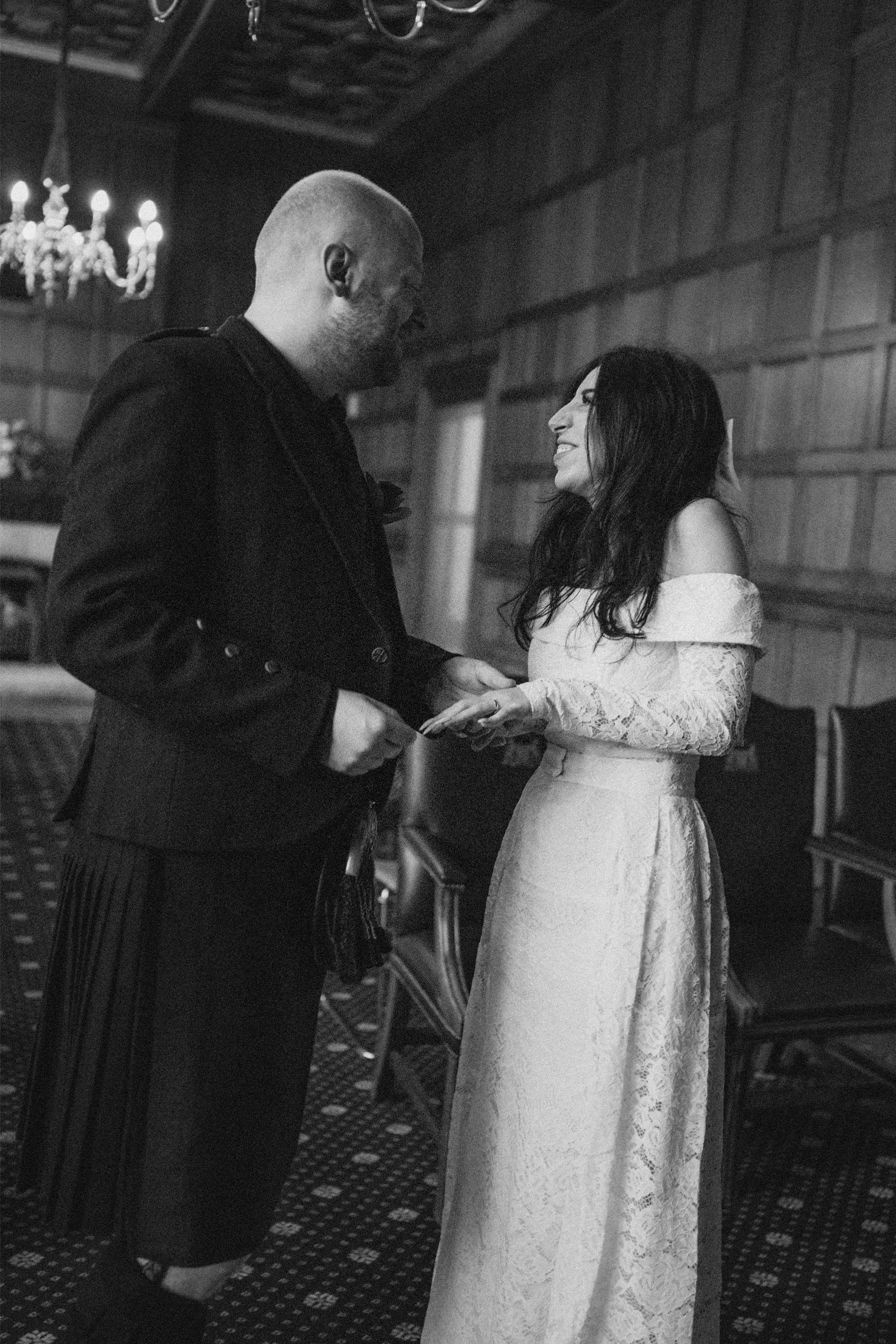 A black and white photo of a couple at their wedding, standing indoors, smiling at each other. The groom is wearing a dark suit with a kilt, and the bride is wearing a long, lace wedding dress.