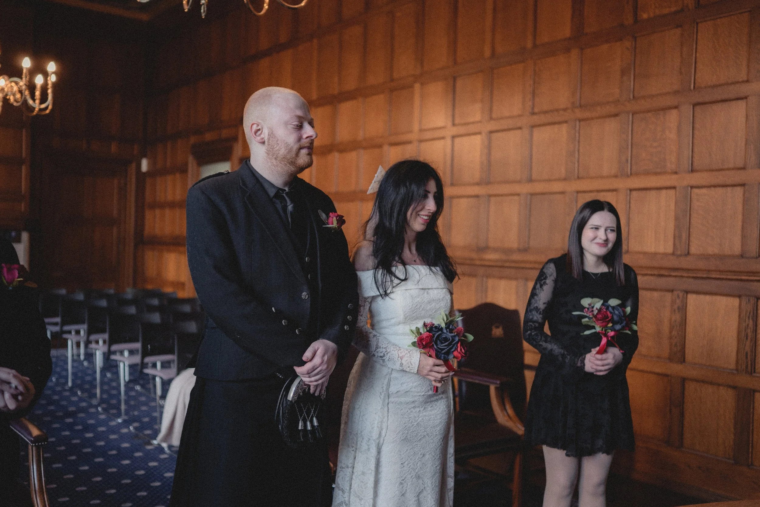 A bride and two bridesmaids standing in a wood-paneled room during a wedding ceremony, holding bouquets, with a chandelier hanging overhead.