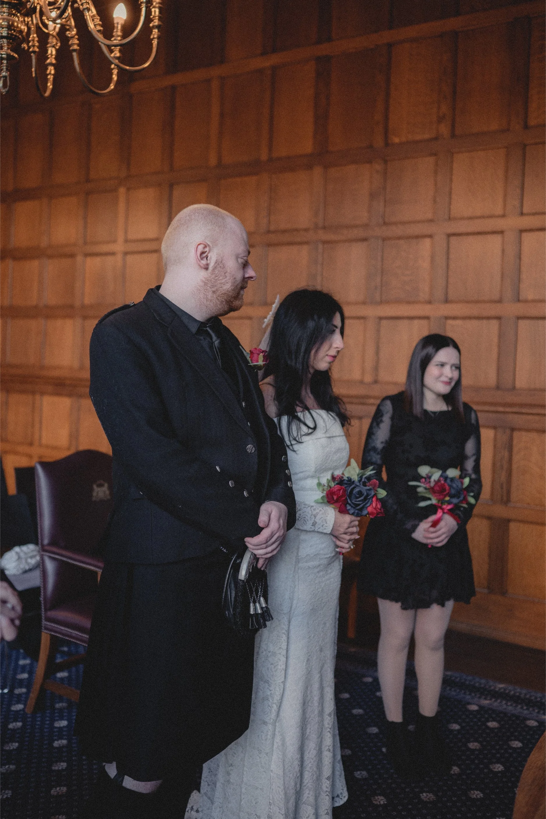 Three people standing in a wood-paneled room attending a wedding ceremony. A man on the left with a shaved head and beard wearing black, a woman in the center in a cream lace dress holding a bouquet of red and dark purple flowers, and another woman o